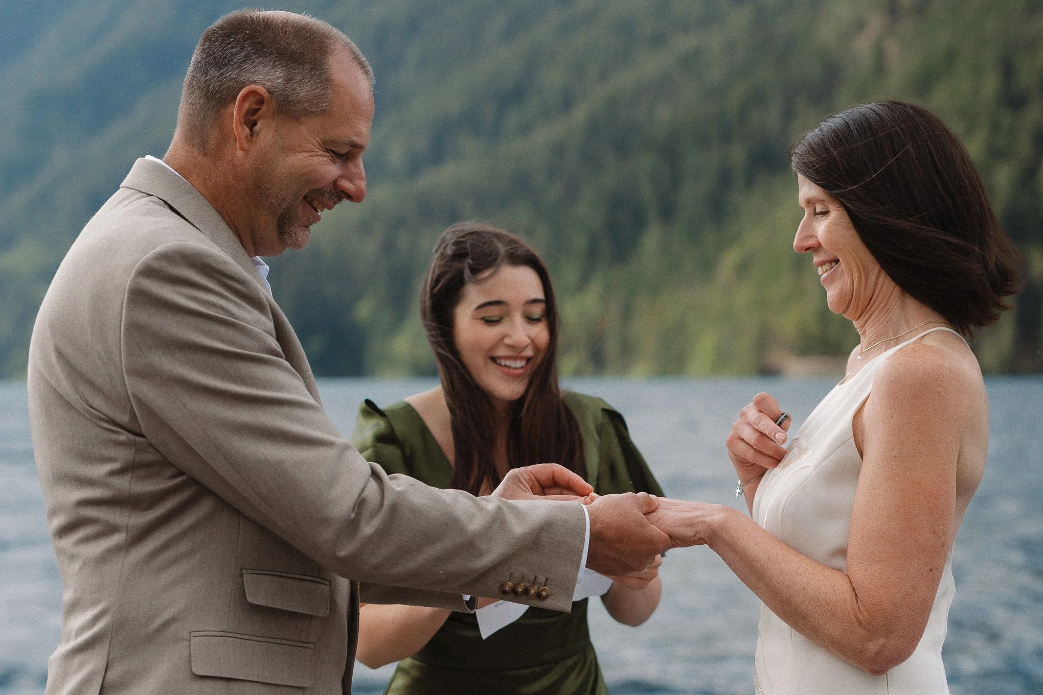 A man and woman exchanging rings during a wedding ceremony outdoors near a lake or river, with a young woman in the background smiling.