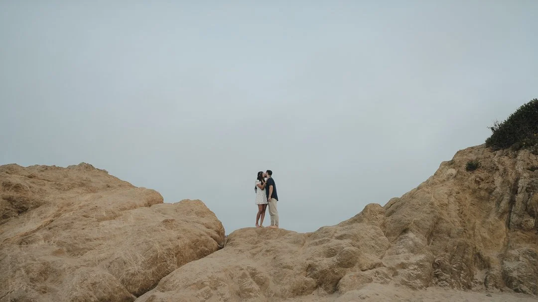 A couple stands together on a rocky cliff, sharing a kiss with a cloudy sky in the background.