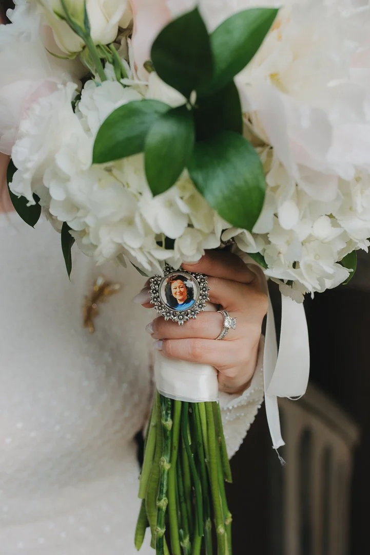A bride holding a bouquet of white flowers with green leaves, featuring a personal photo ring among the flowers.