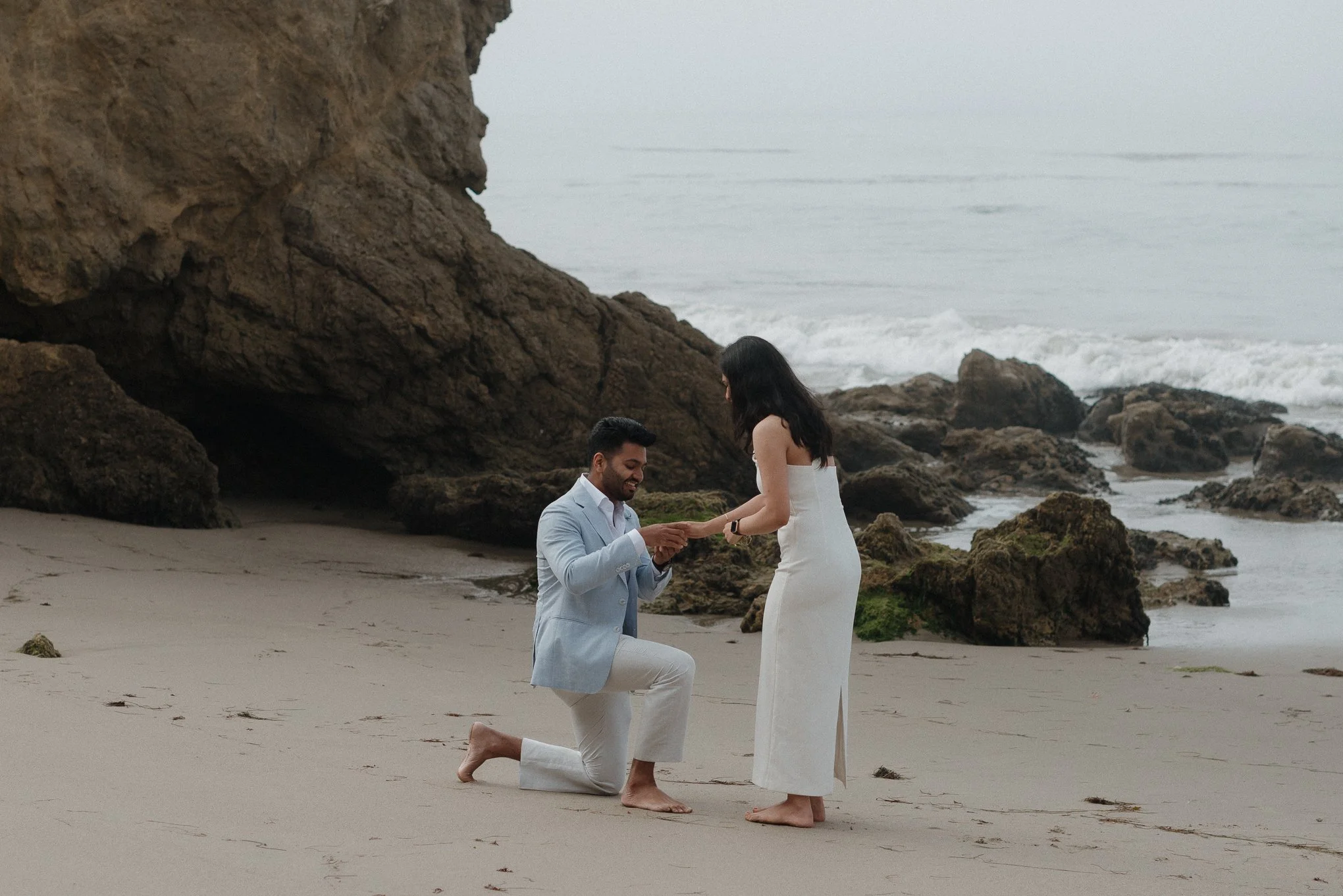 A man in a light blue suit proposes marriage to a woman in a white dress on a sandy beach with rocky cliffs and ocean waves in the background.