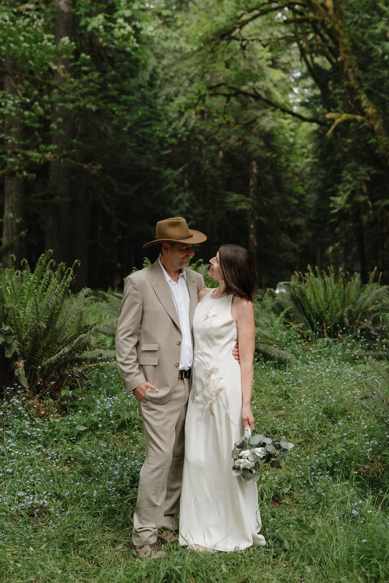A man and woman standing close together outdoors in a lush green forest, gazing at each other, with the woman holding a bouquet of white flowers.