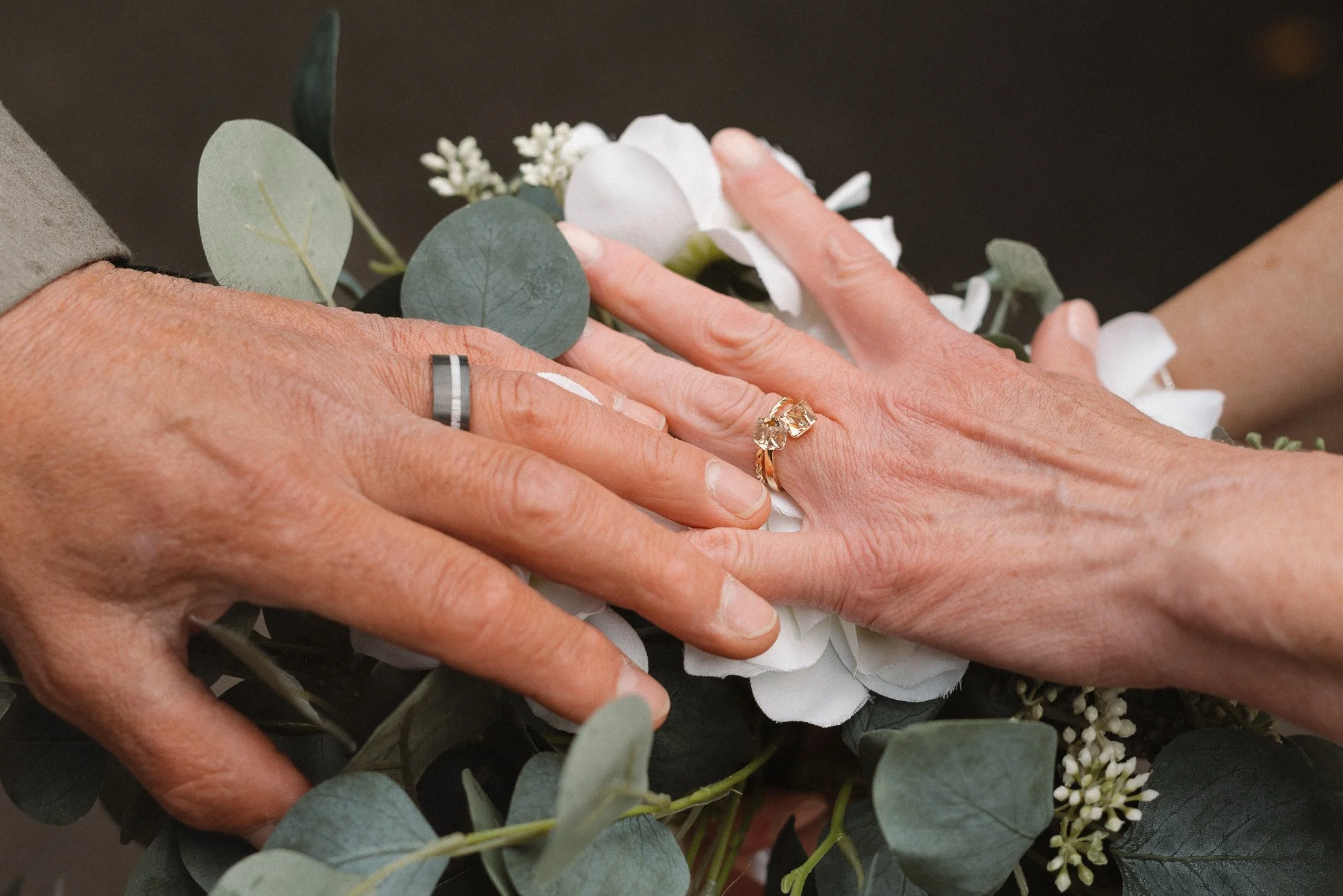 A person's hands with wedding rings are resting on white and green floral arrangement, including white flowers and eucalyptus leaves.