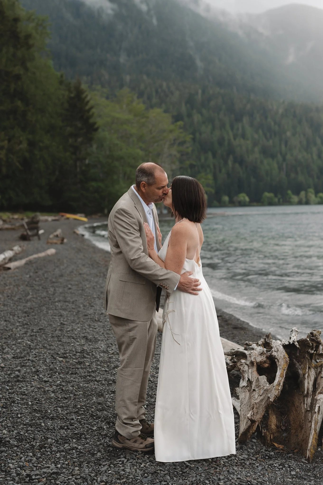 A couple in wedding attire sharing a kiss on a rocky beach with a forested mountain in the background.