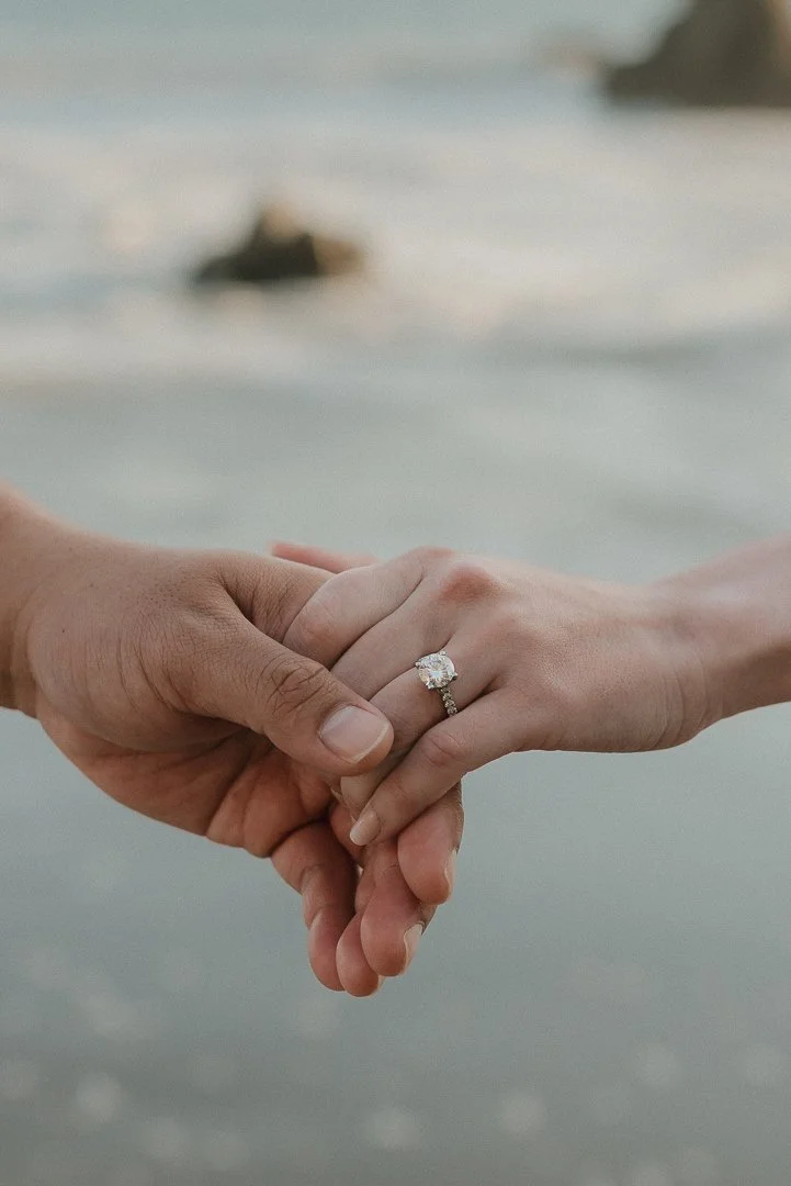 Two people holding hands, with a close-up focus on an engagement ring on the woman's finger, against a blurred beach background with rocks in the distance.