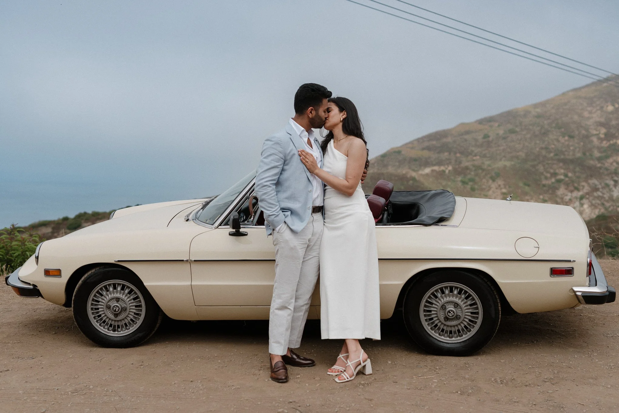 A man and woman kissing on top of a vintage beige convertible car in a scenic outdoor location with hills and power lines in the background.
