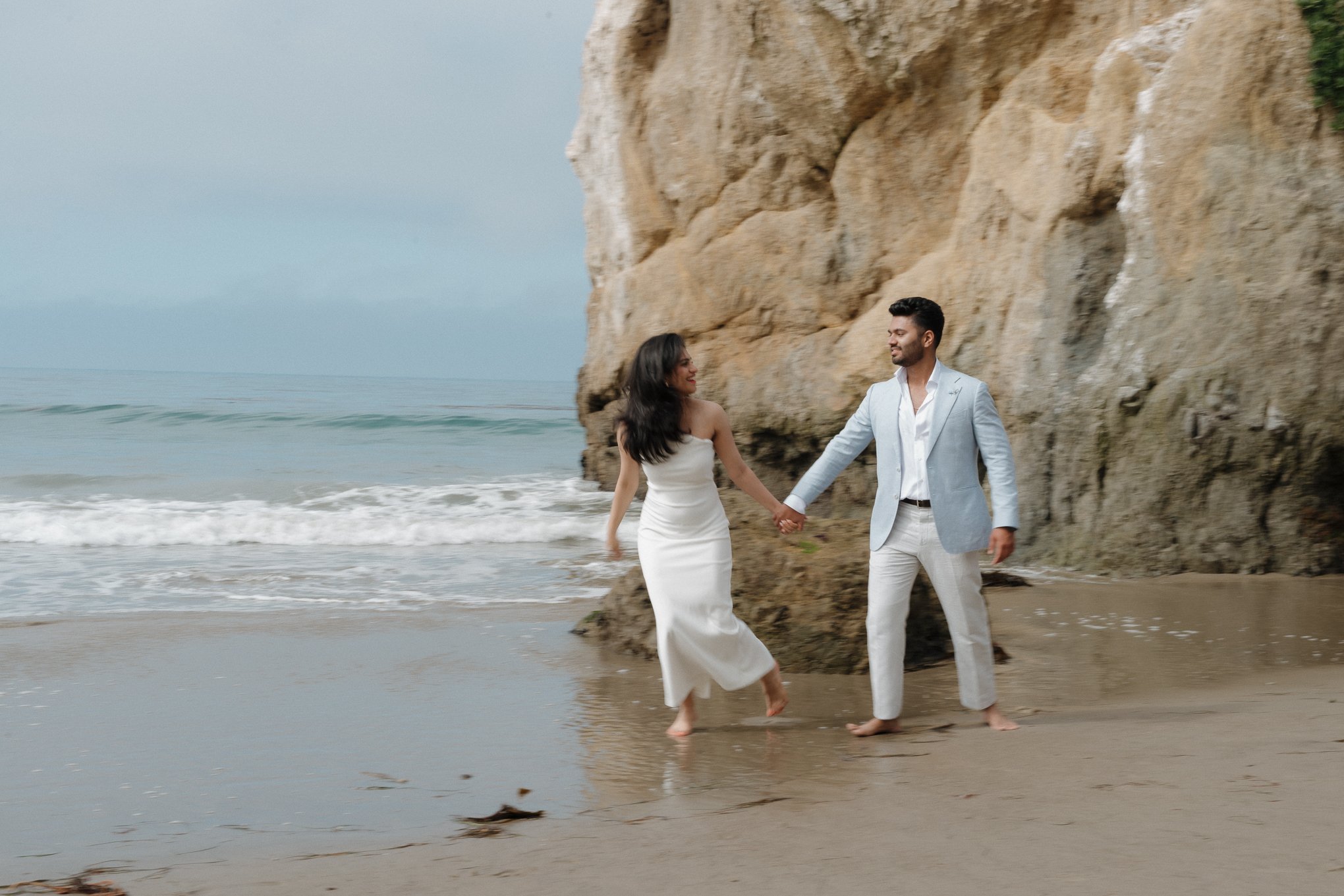 A couple dressed in wedding attire walking hand in hand on the beach near a large rock formation, with the ocean in the background.