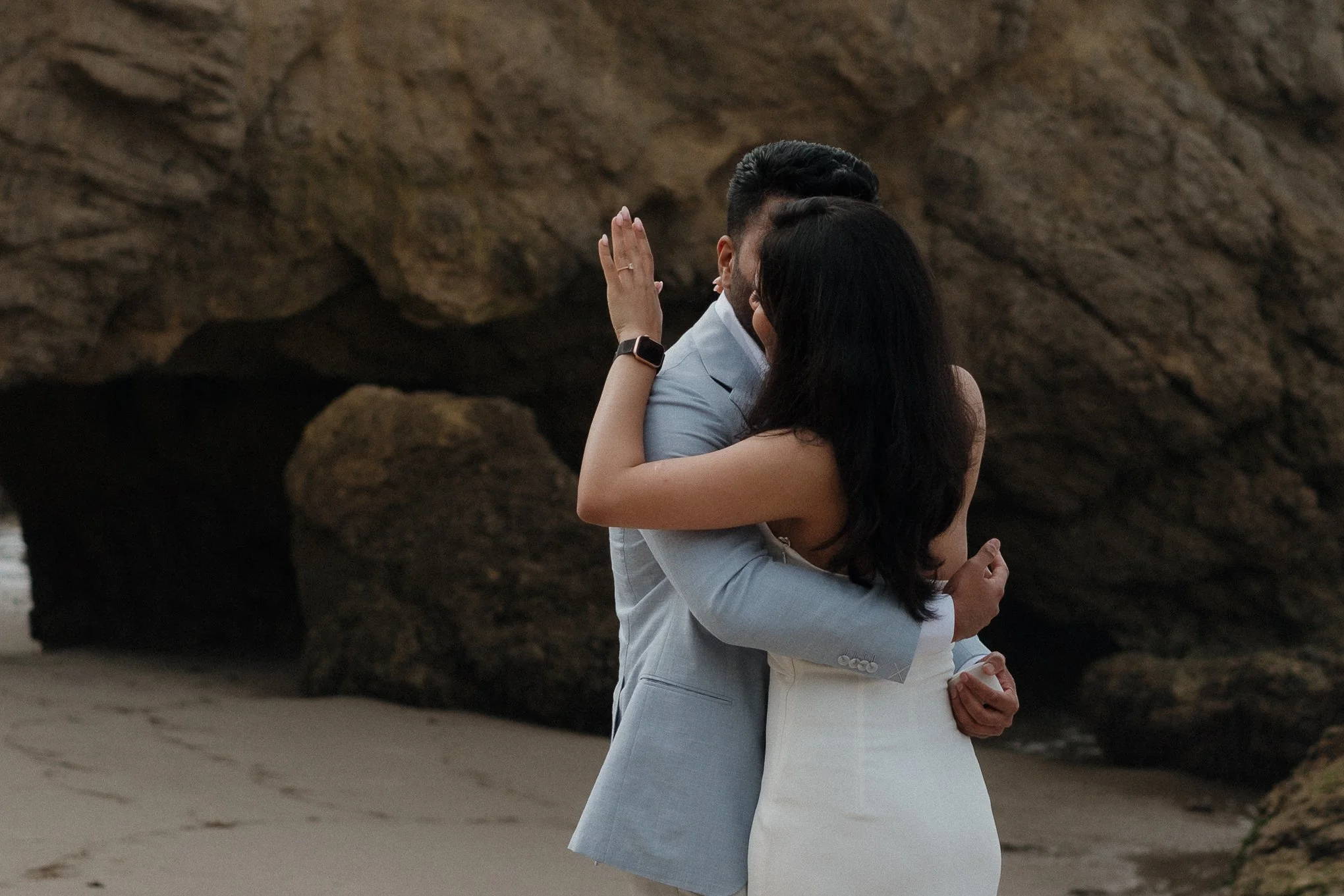A couple embracing on the beach, with large rocks in the background, the woman has long dark hair and is wearing a white dress, and the man has short dark hair and is dressed in a light-colored suit.