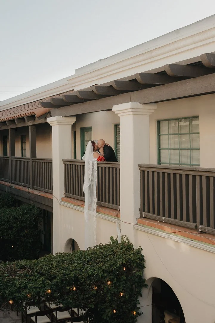 A bride and groom sharing a kiss on a balcony of a white, two-story building with green window frames and a dark wooden railing. The bride's veil is hanging down and a bouquet of red flowers is visible.