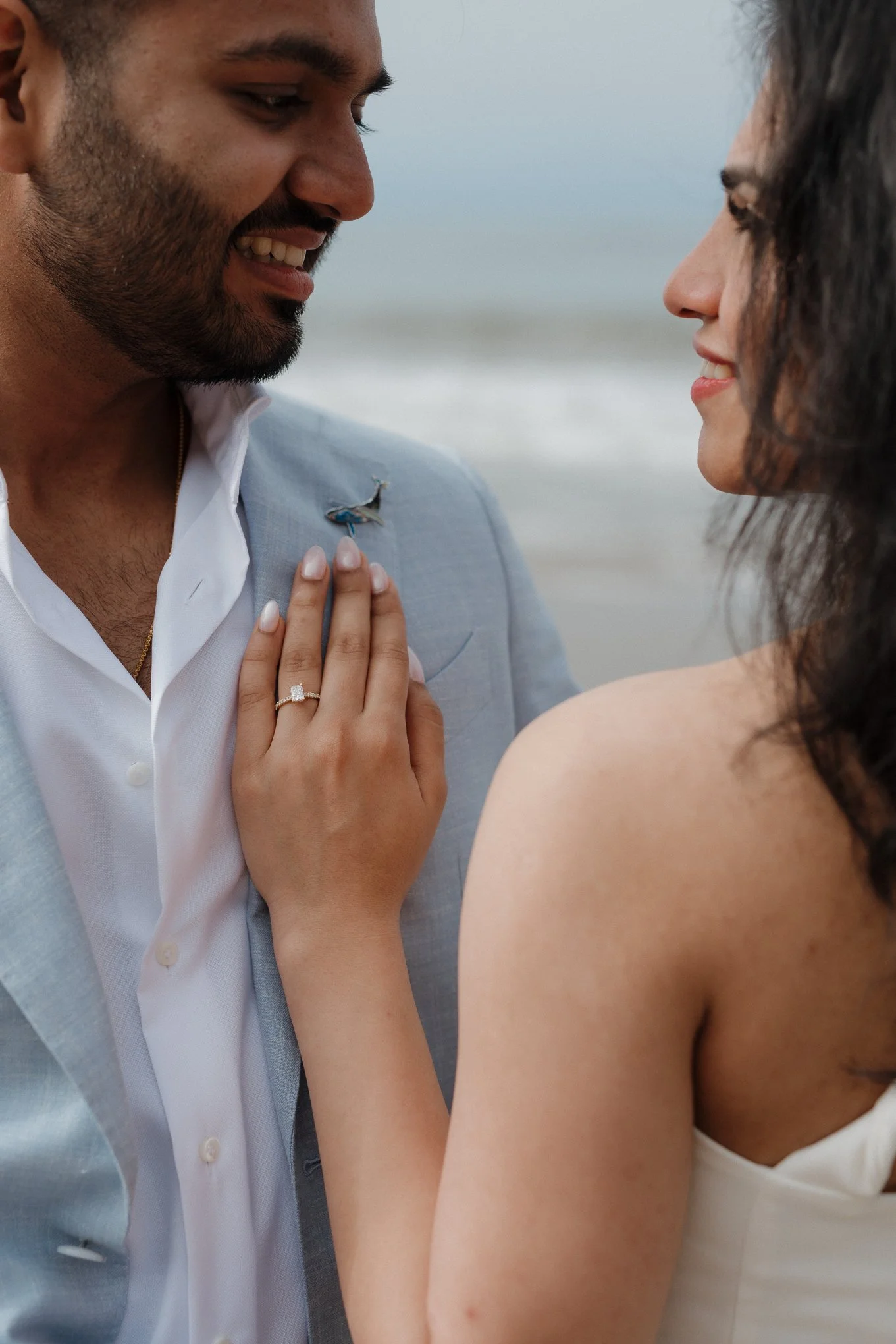 A man and a woman face each other close at the beach, smiling. The woman has her hand on the man's chest, showing an engagement ring.