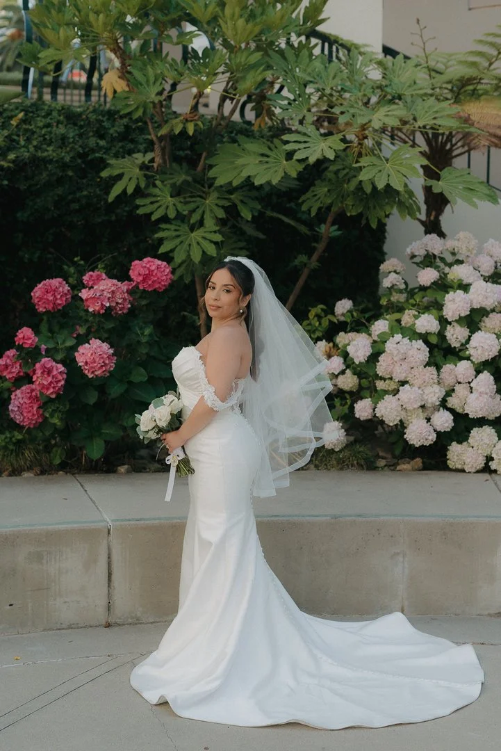 A bride in a white wedding gown holding a bouquet, standing outdoors in front of pink and white hydrangea bushes and green trees.