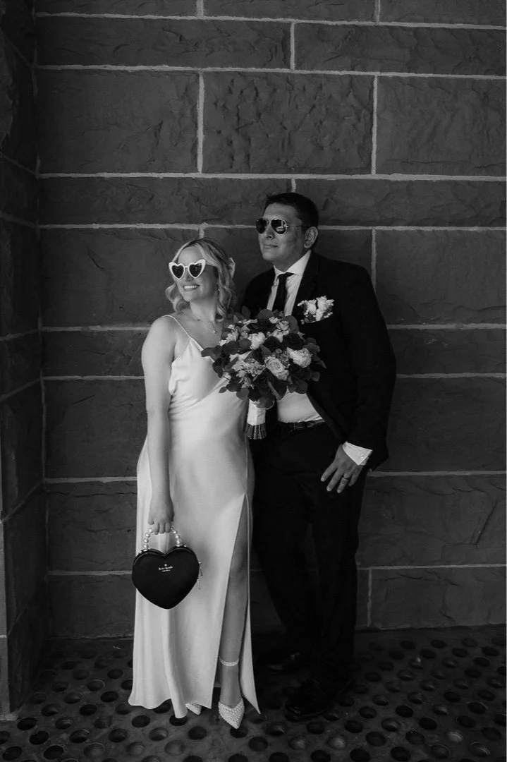 A black and white photo of a couple dressed for a wedding, standing against a brick wall. The woman is wearing a satin wedding dress, heart-shaped sunglasses, and holding a heart-shaped purse. The man is dressed in a suit with sunglasses and is holding a bouquet of flowers.