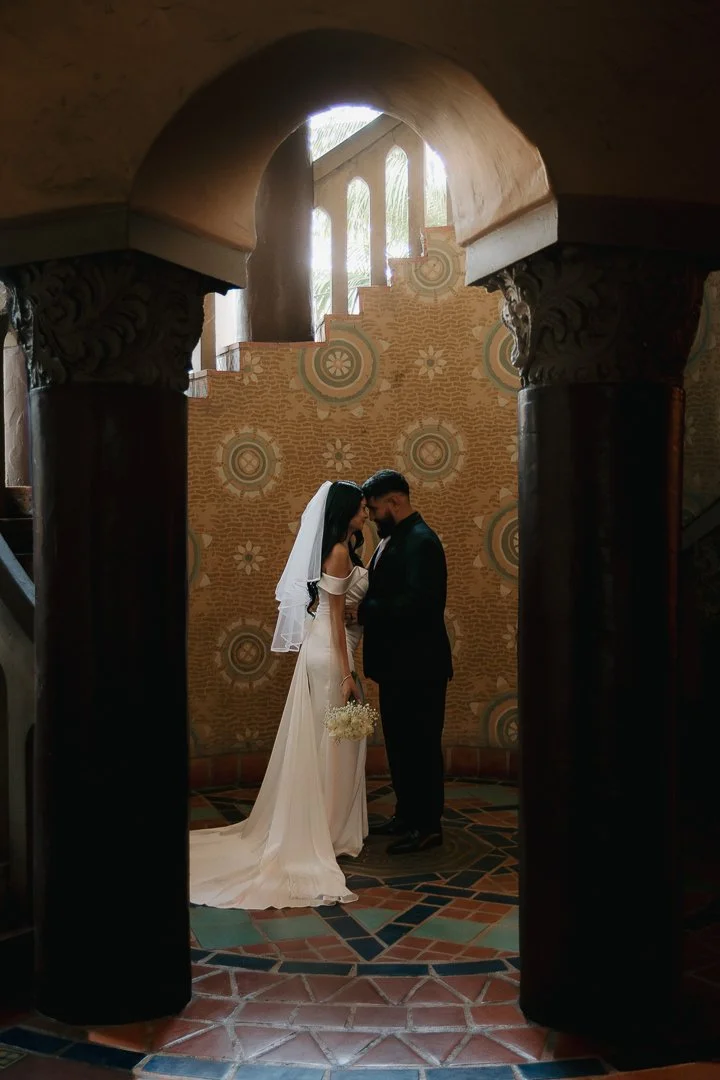A bride and groom in wedding attire standing close, touching foreheads and holding hands inside a decorative, arched interior with patterned walls and sunlight streaming through windows.
