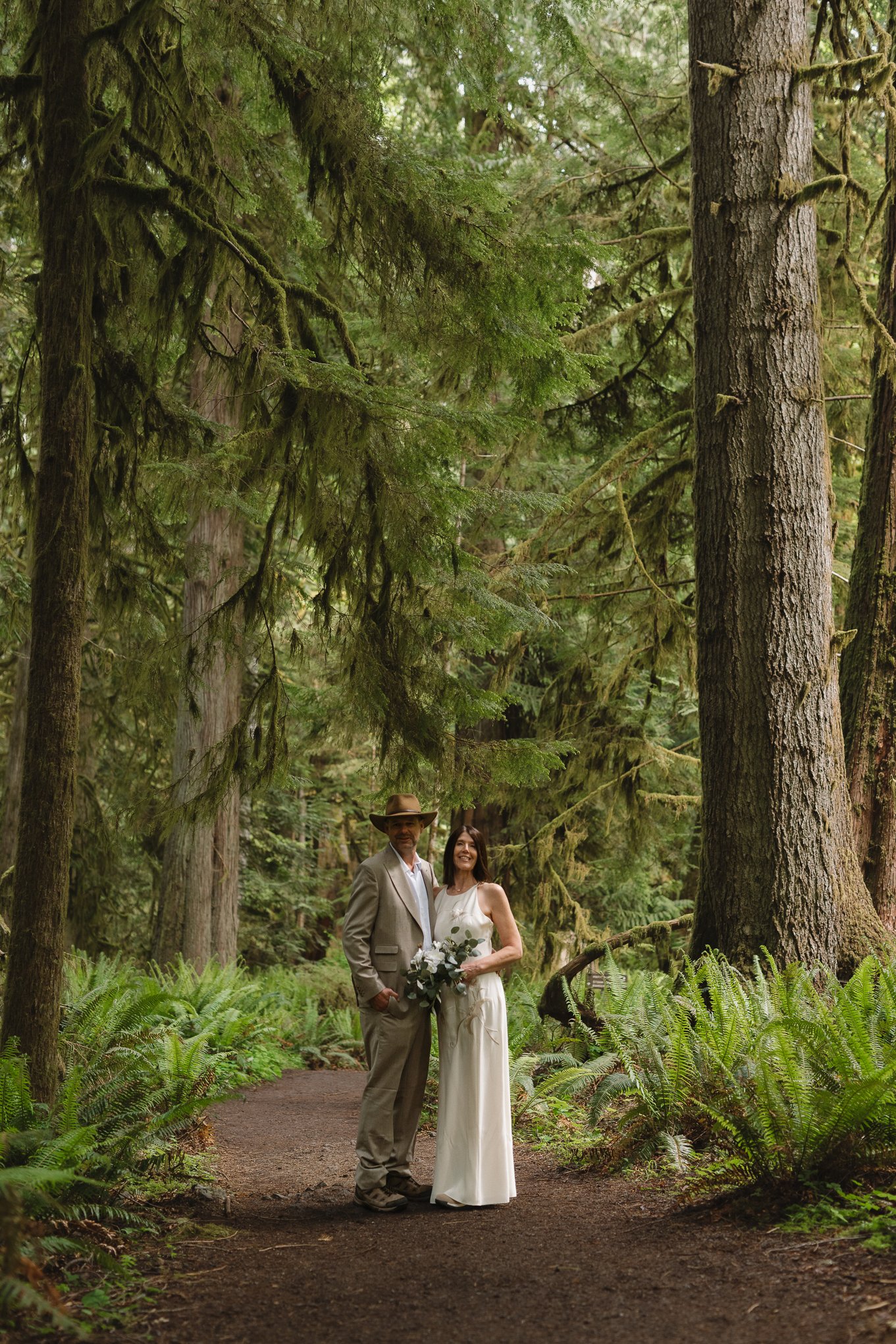 A couple in wedding attire standing on a forest trail surrounded by lush green ferns and tall trees, some with moss, in a wooded area.