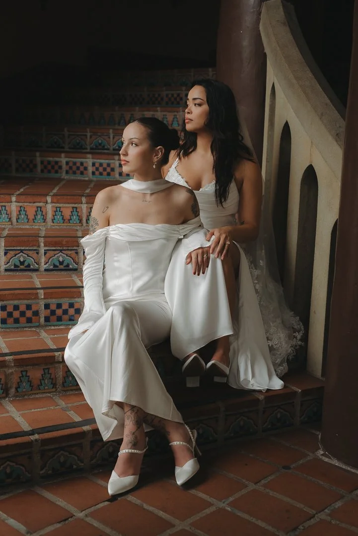 Two women in white dresses sitting on decorative tile stairs, one with dark hair and the other with light hair, in what appears to be a historical or elegant setting.