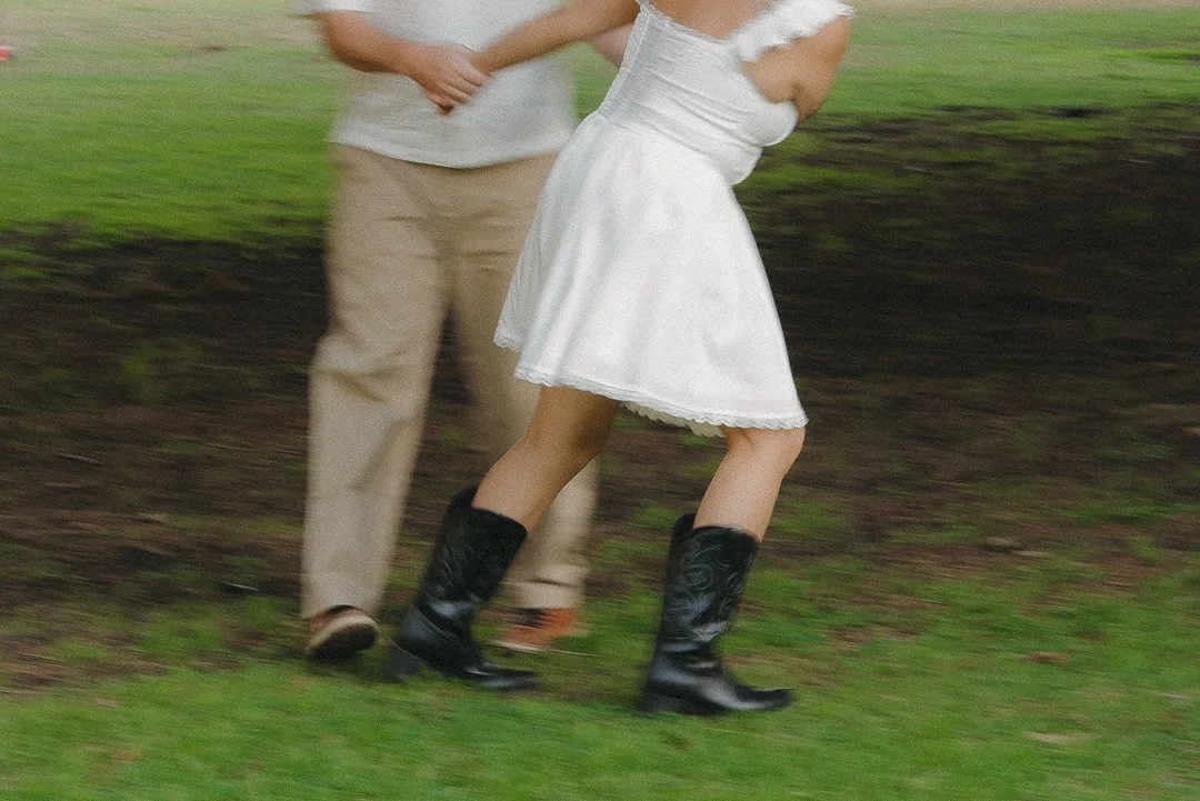 Partially visible people walking on grass, woman in white dress and black boots, man in beige pants and white shirt.