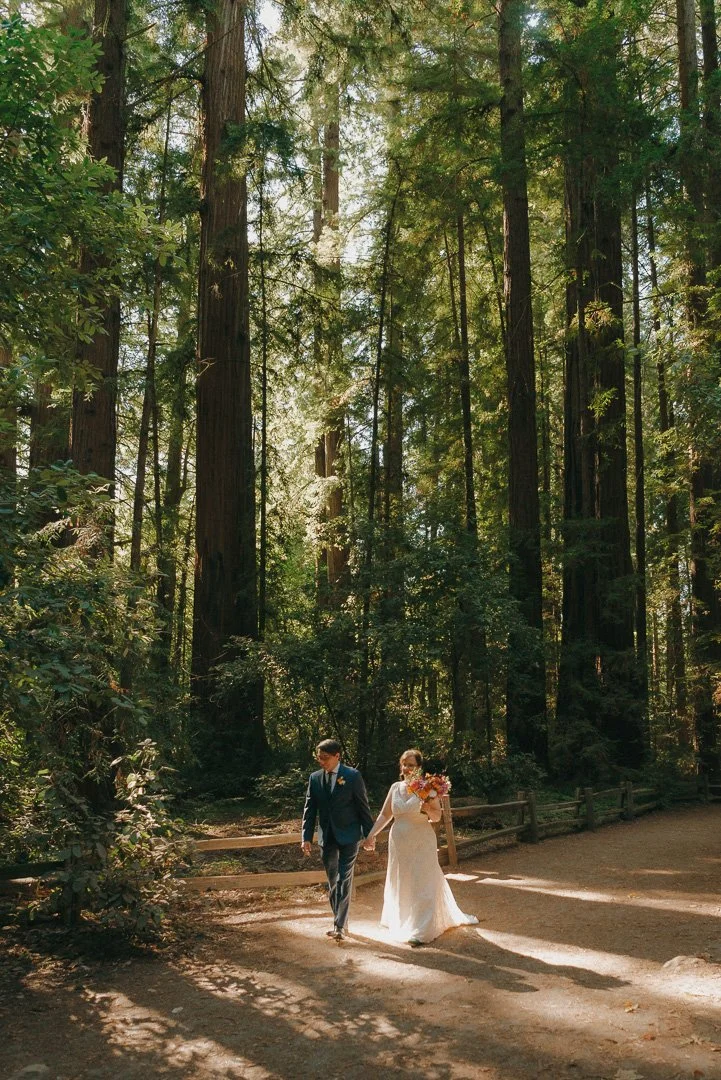 A bride and groom walking hand in hand along a forest trail, sunlight filtering through tall trees, with the bride holding a bouquet of flowers.
