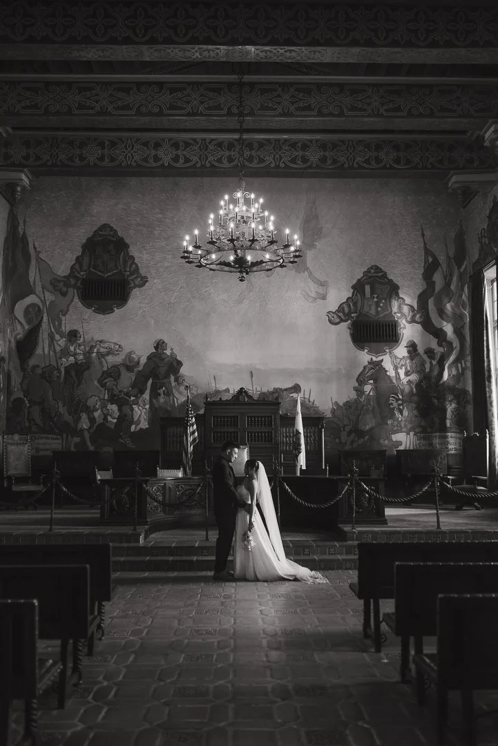 A bride and groom stand together in a historic courtroom with a mural on the wall behind them. The room features an ornate chandelier hanging from the ceiling, several flags, and wooden benches.