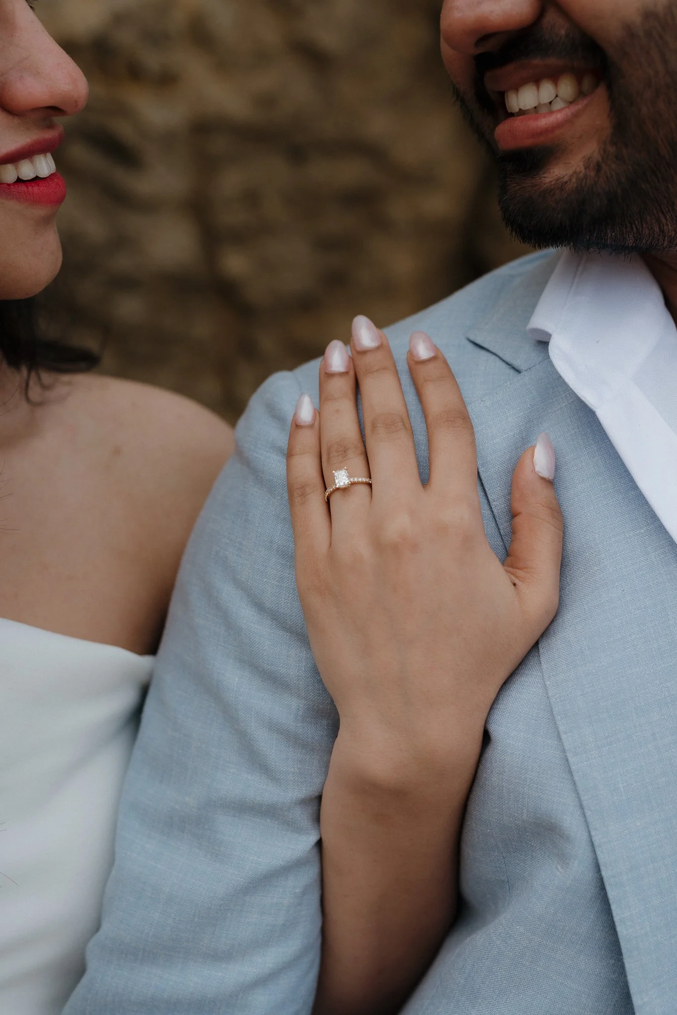 A close-up of a smiling couple, focusing on their faces and the woman's hand with an engagement ring resting on the man's shoulder. The woman has red lipstick and well-manicured nails, and the man has a beard and is wearing a light blue blazer.