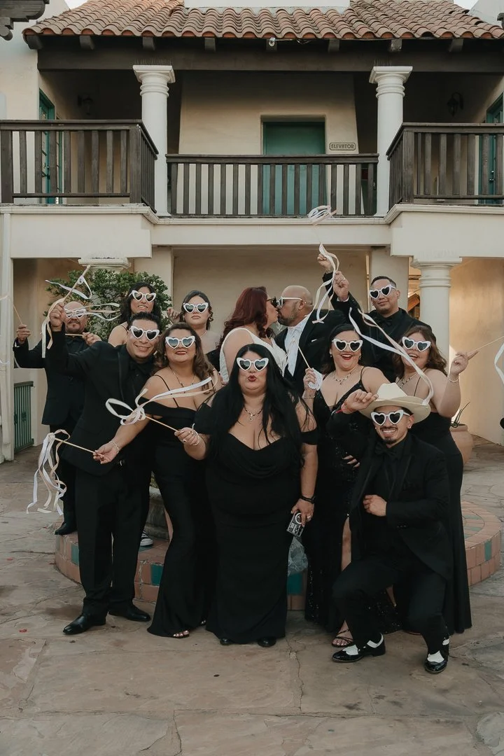 Group of people dressed in black, wearing heart-shaped sunglasses, holding white ribbons, posing together outside a two-story house with balconies.