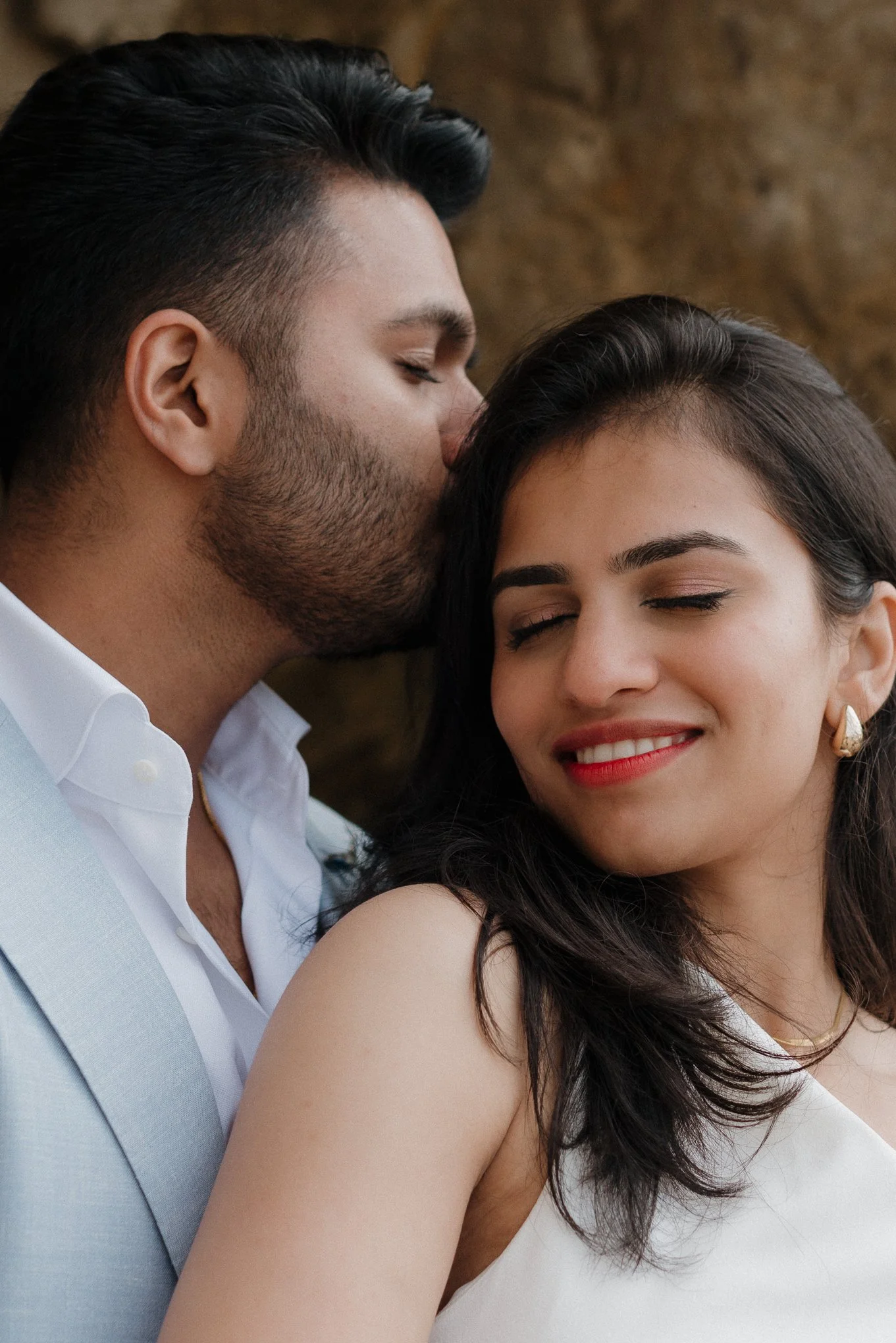 A close-up of a couple with closed eyes, the man giving a gentle kiss on the woman's forehead, who is smiling with dark hair and red lipstick, against a brown brick background.