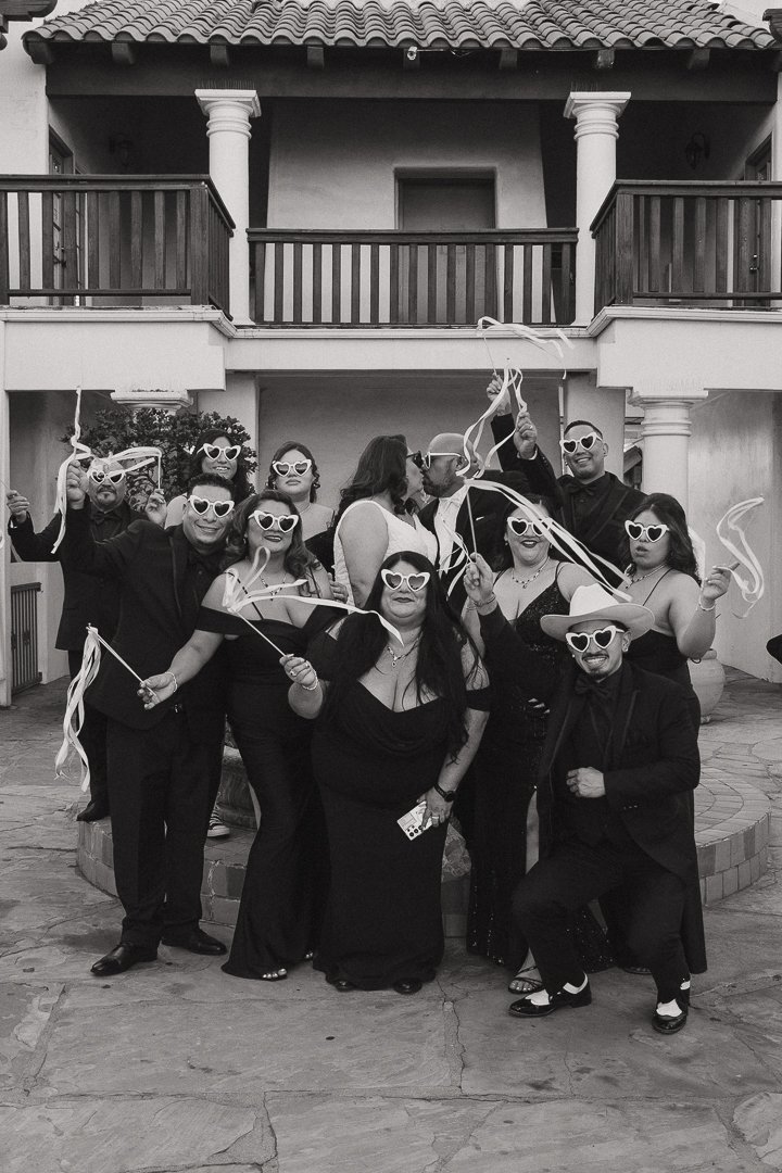 A group of people celebrating outdoors in front of a house, wearing heart-shaped sunglasses, holding streamers, and dressed in formal black attire, with some wearing hats.