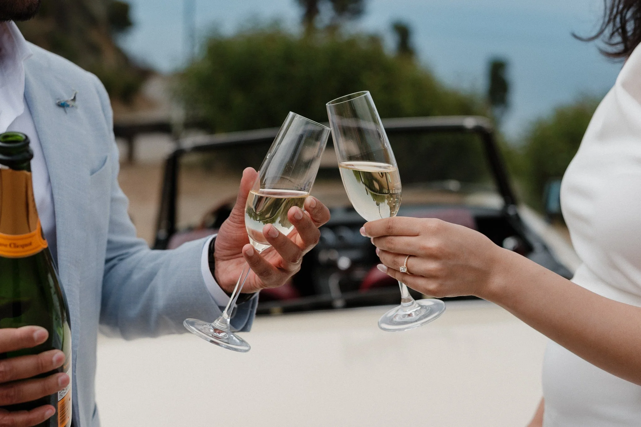 Two people clinking champagne glasses outdoors, with one person holding a bottle of champagne.