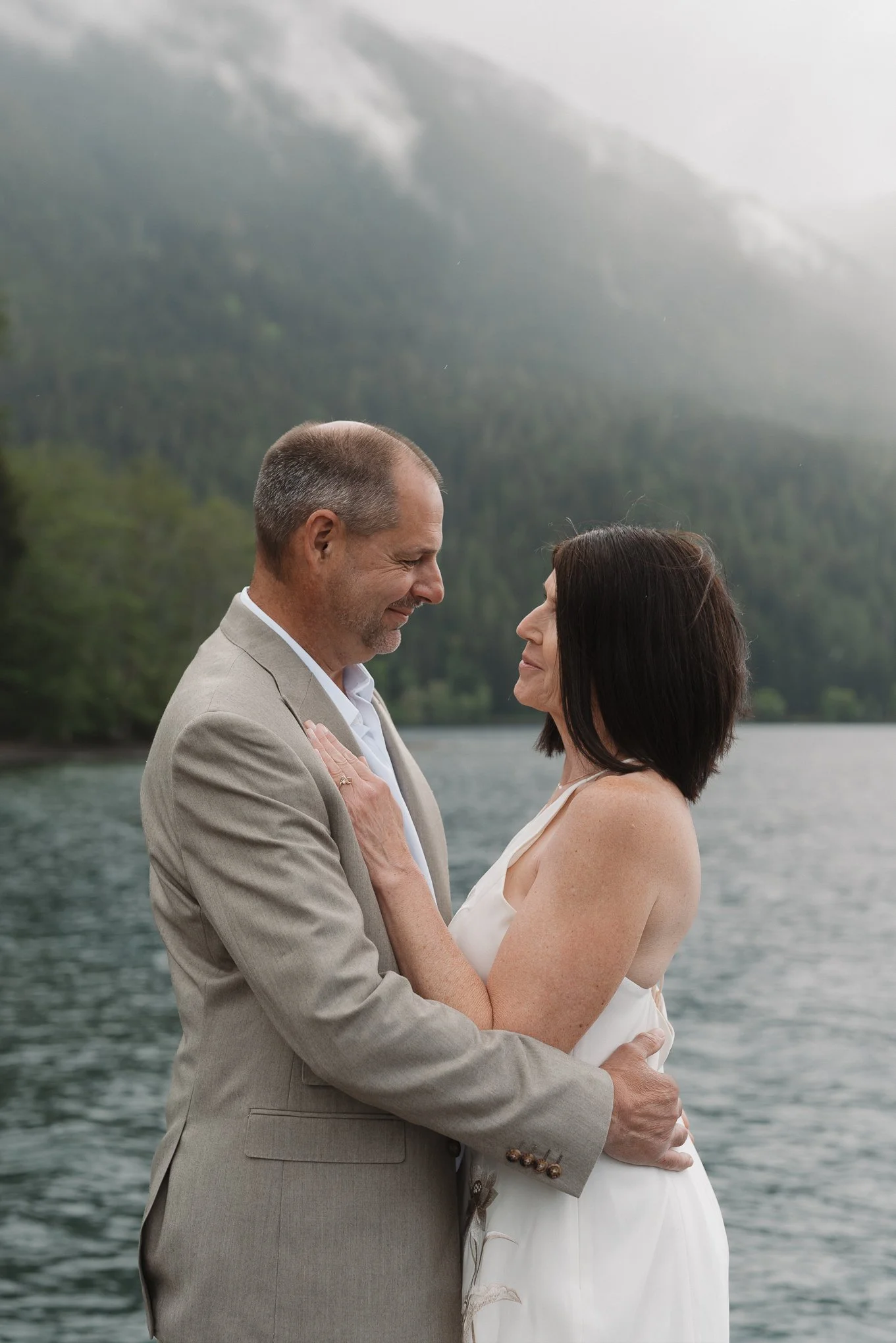 A couple stands face to face outdoors by a body of water with a misty mountain landscape in the background, holding each other and gazing into each other's eyes.