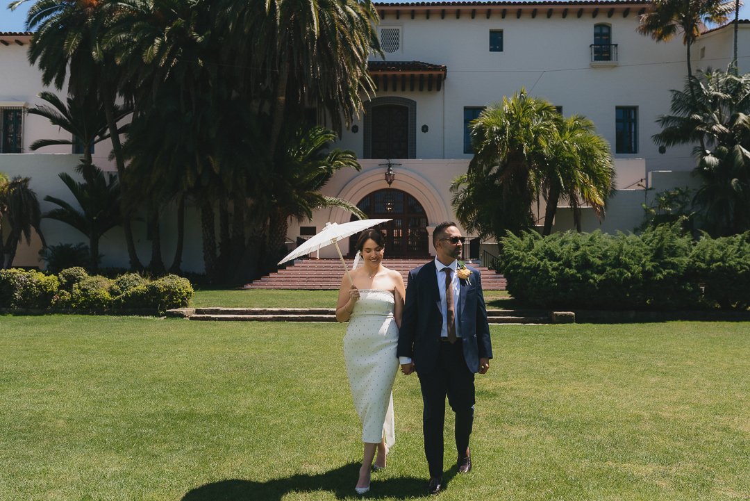A bride and groom walking hand in hand on a lush lawn in front of a white mansion with lush greenery and tall palm trees, the bride holding a white parasol.