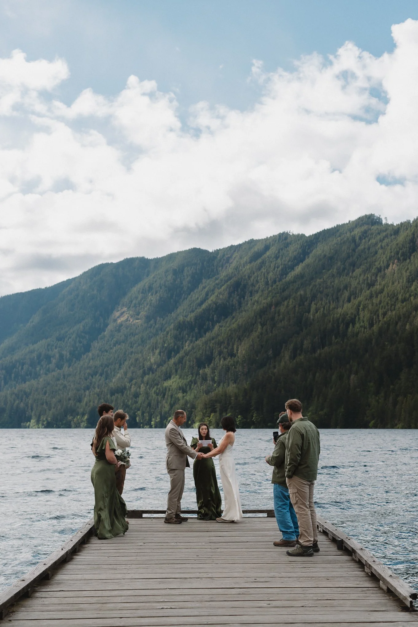 A couple is getting married on a wooden dock in front of a lake surrounded by mountains, with friends and family participating in the ceremony.