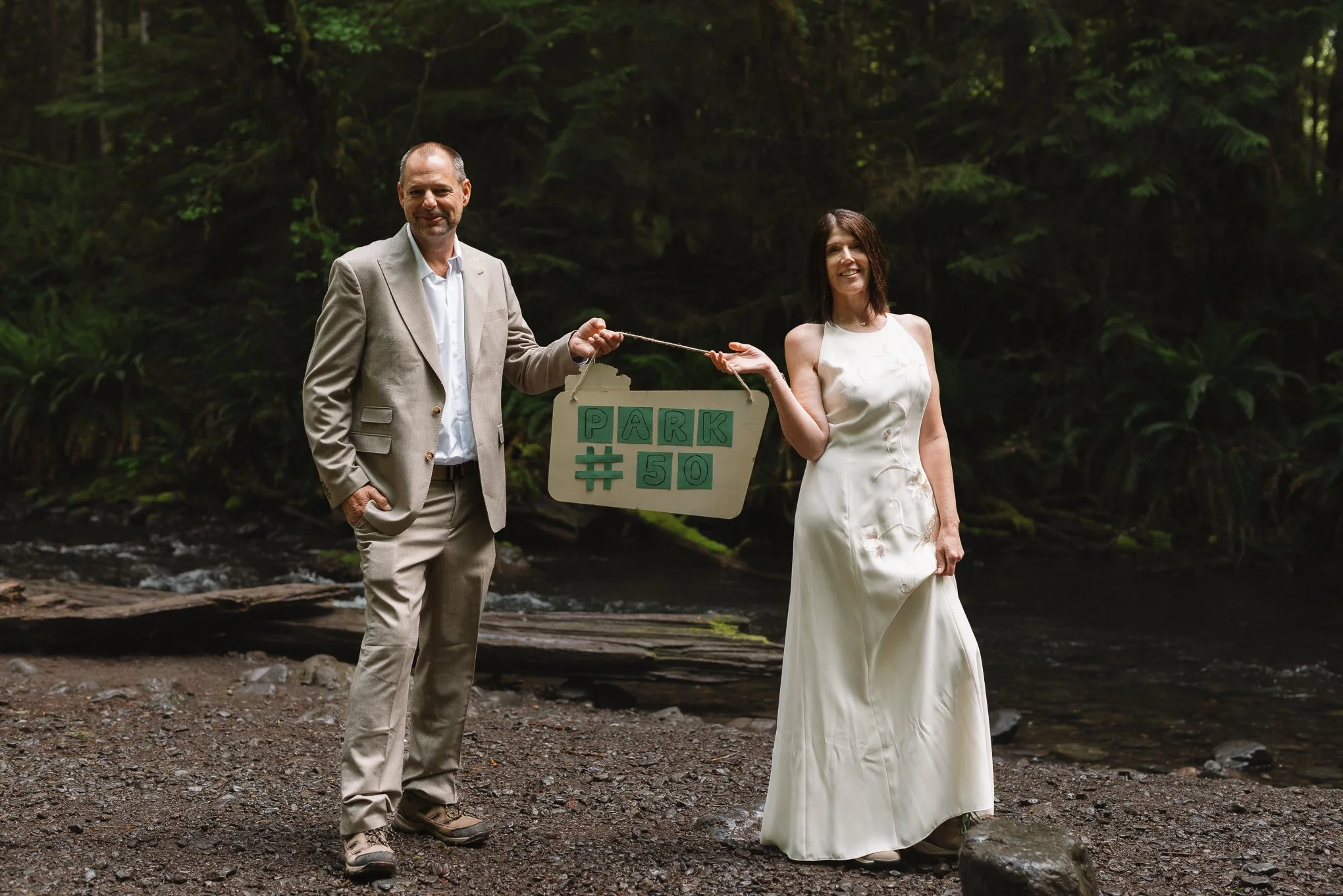 A man in a beige suit holding a sign that says 'PARK #50' and a woman in a white dress standing outdoors near a stream in a wooded area.