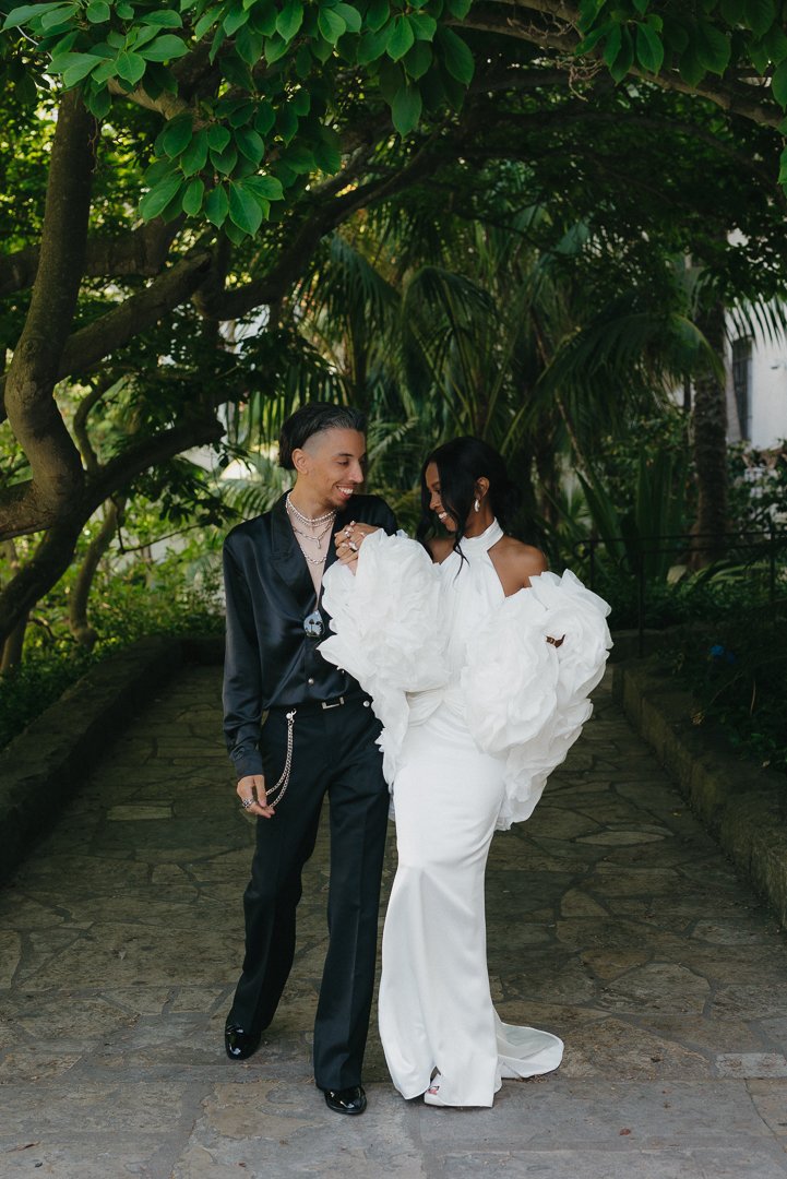 A couple dressed in stylish formal attire standing on a stone pathway surrounded by lush greenery, smiling and holding hands.