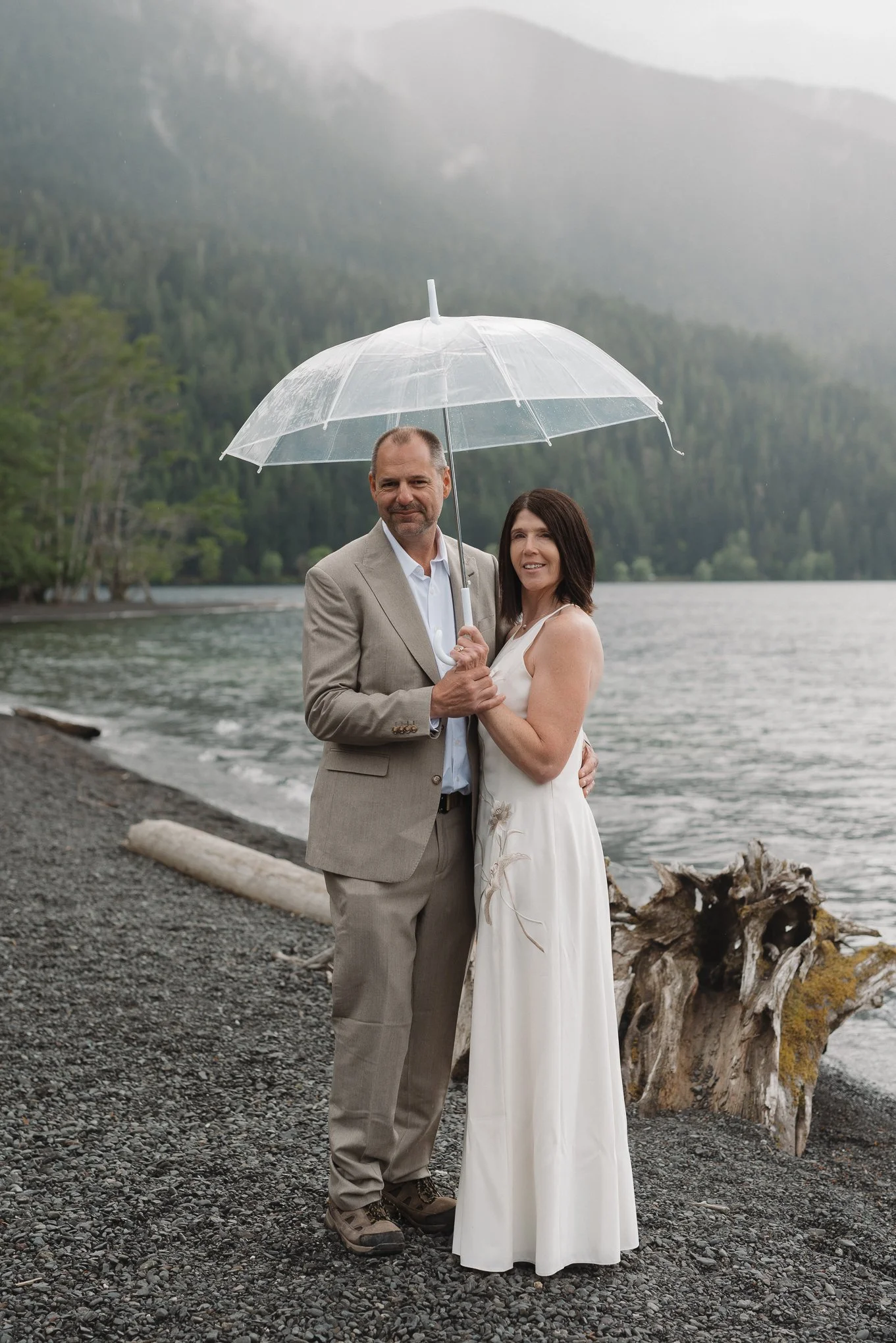 A couple is standing by a lake on a pebbled shore, holding transparent umbrellas, with a mountain range and trees in the background. The man is in a beige suit and the woman is in a white dress.