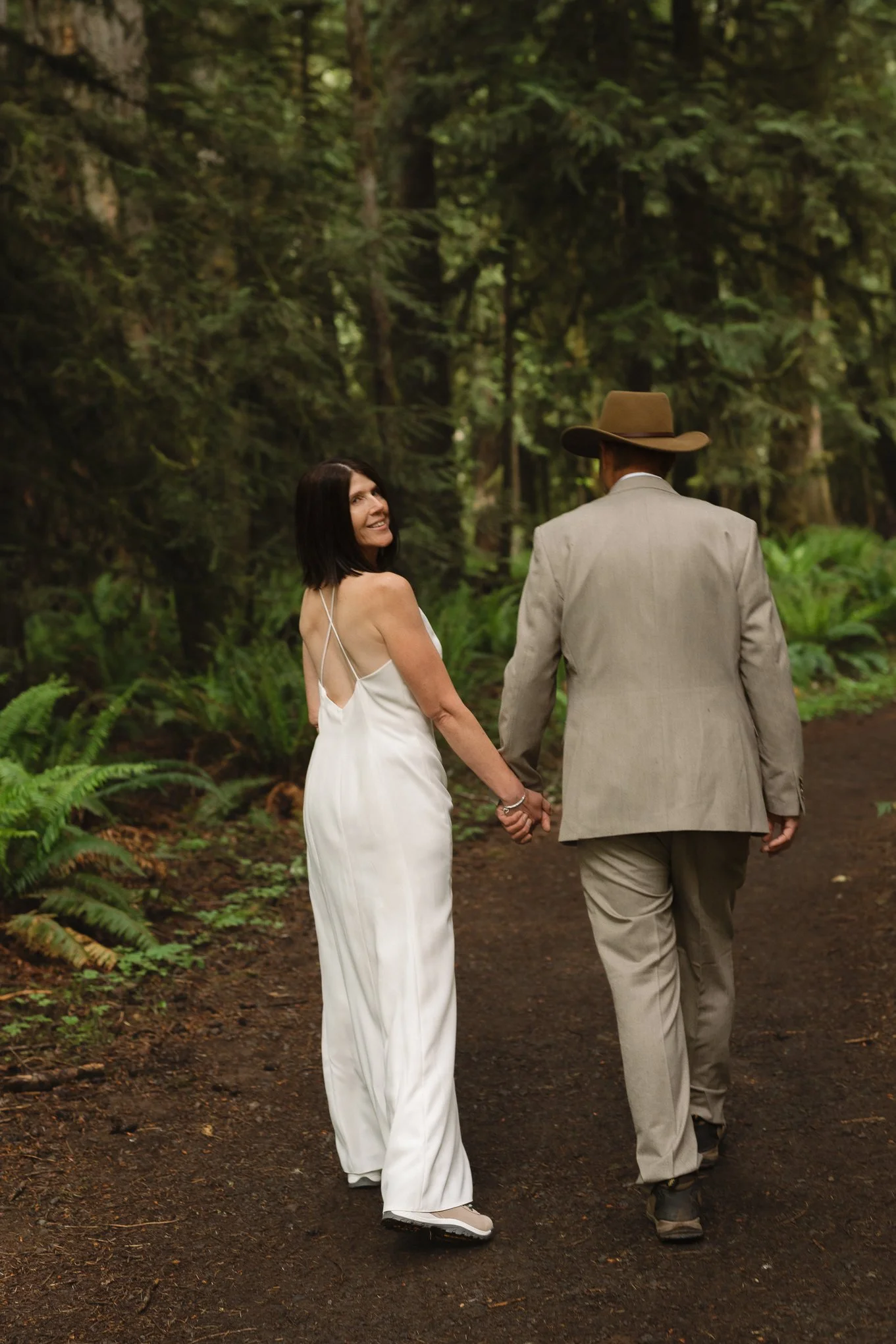 A woman in a white dress holding hands with a man in a beige suit and large brown hat, walking through a lush forest.