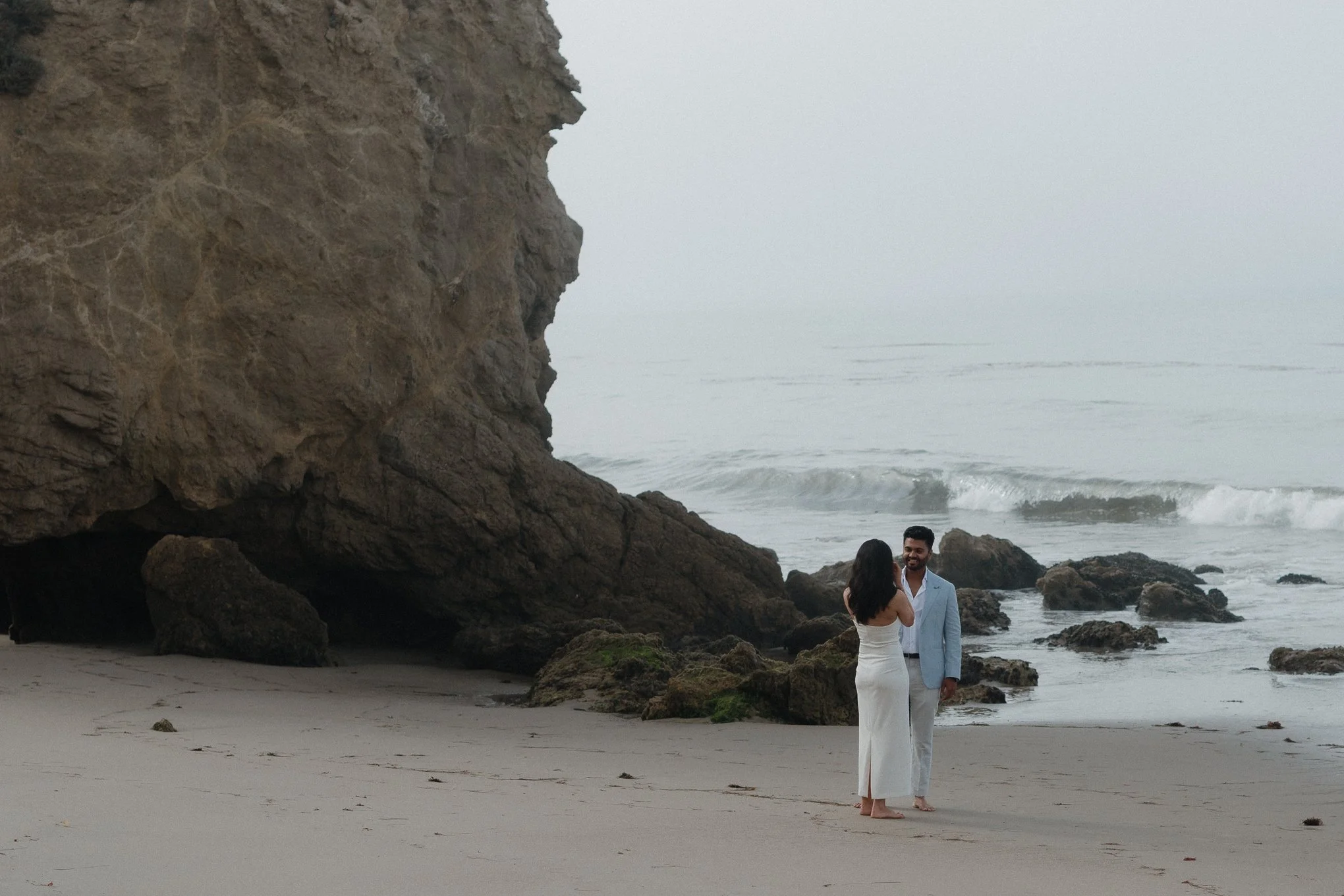 A couple dressed in formal wedding attire standing on a beach near rocks and the ocean, with a large cliff on the left side, during overcast weather.