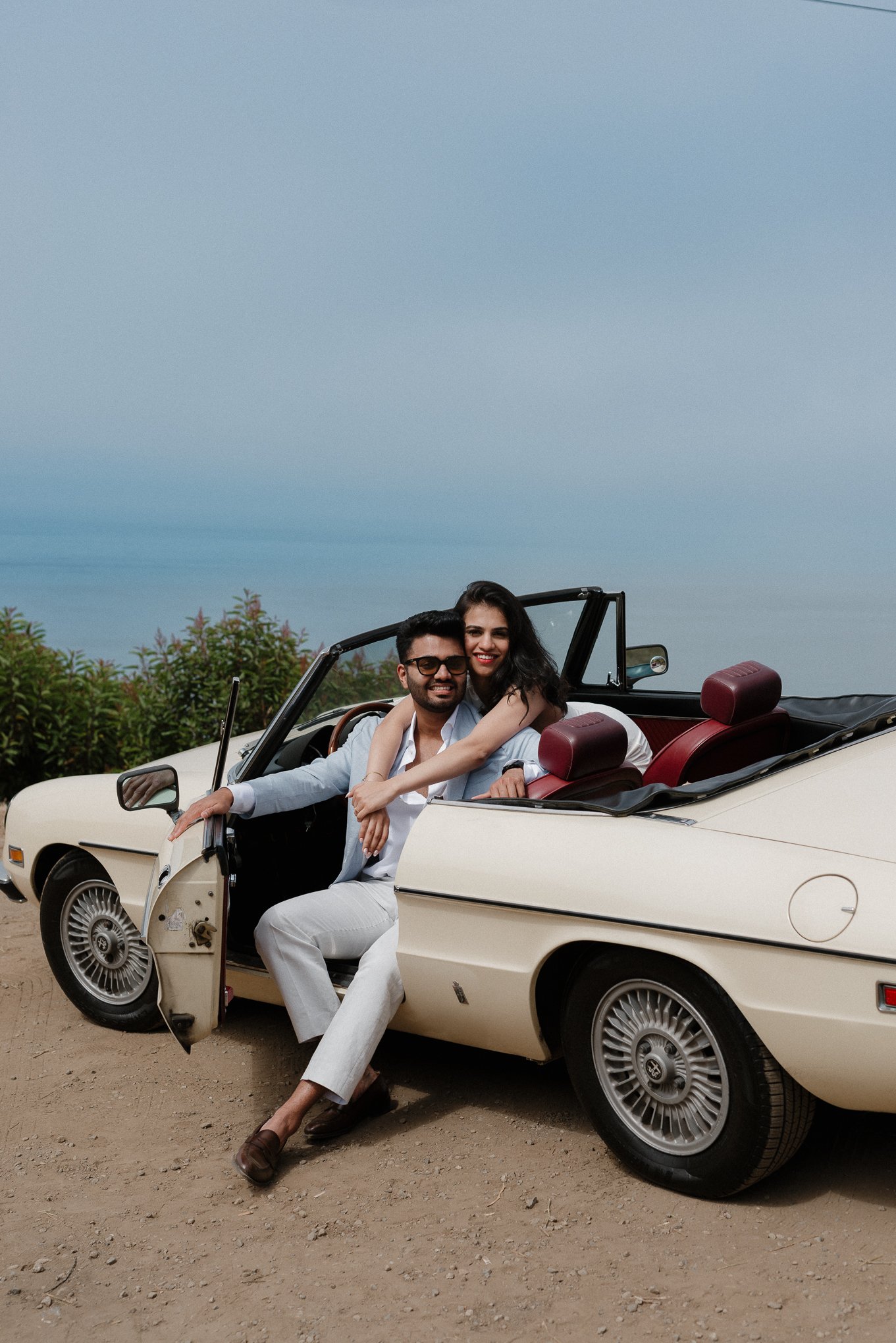 A couple sitting in a vintage white convertible car, outdoors on a dirt ground, with greenery and a cloudy sky in the background. The man wears sunglasses and a white blazer, and the woman is smiling, hugging the man from behind.