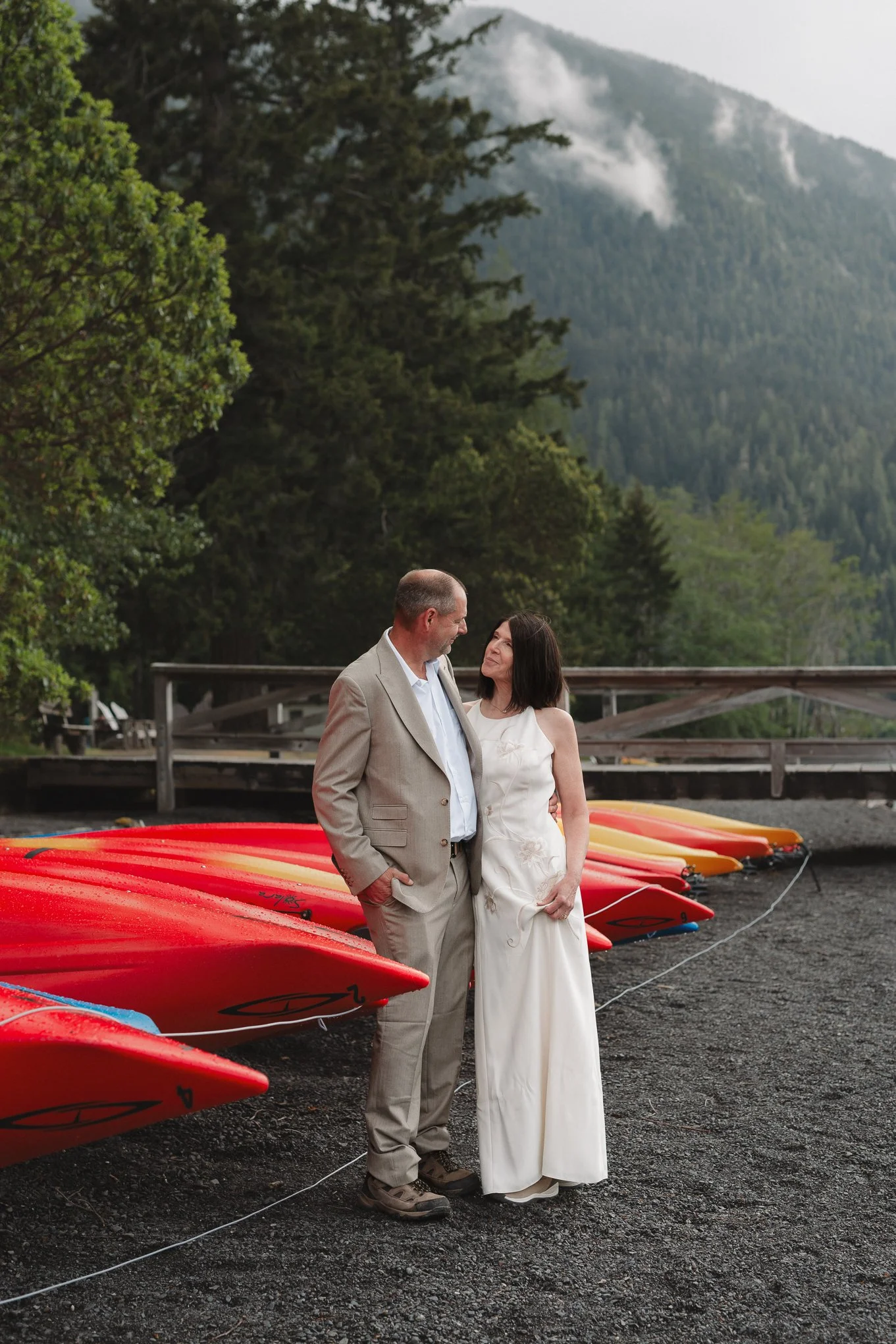 A man and woman standing close together by a row of red and yellow kayaks on a dock, overlooking a scenic mountain landscape with lush green trees and a cloudy sky.