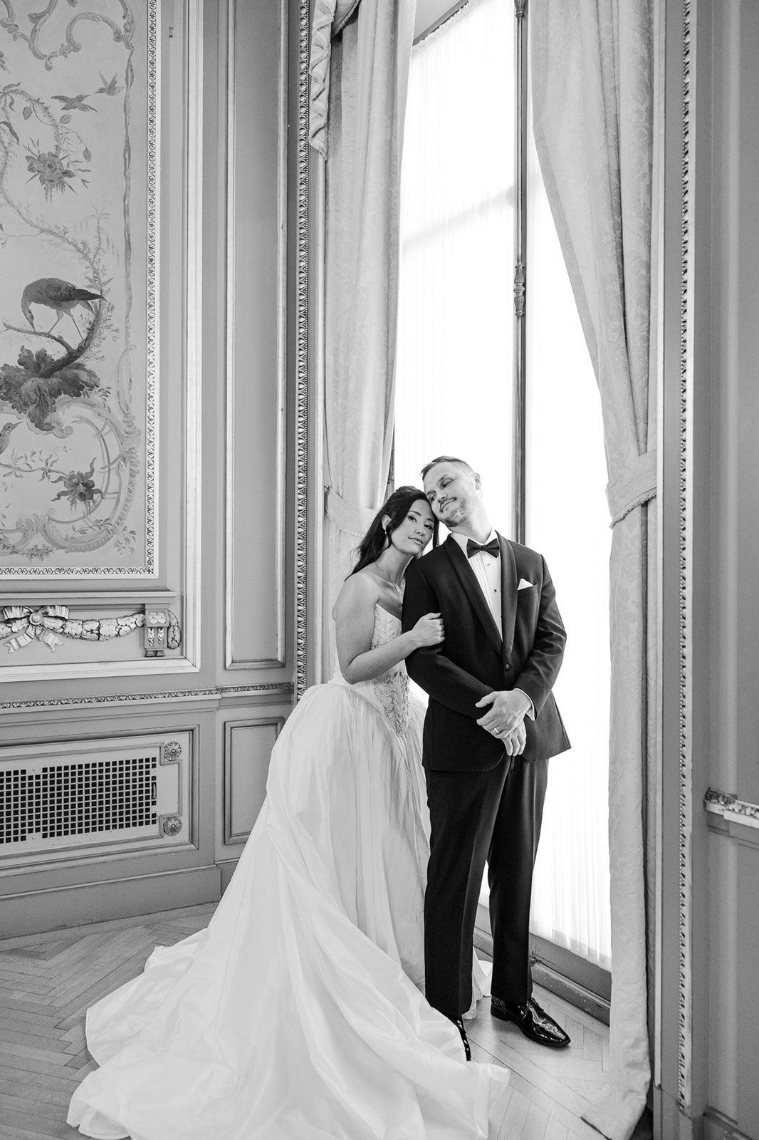 Black and white photo of a bride and groom standing together near a large window in an ornate room, with the bride gently leaning her head on the groom's shoulder.