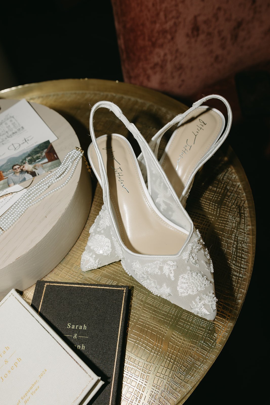 A pair of white, lace embellished high-heeled wedding shoes on a golden tray, along with photo books, a pearl hairband, and a wedding card.