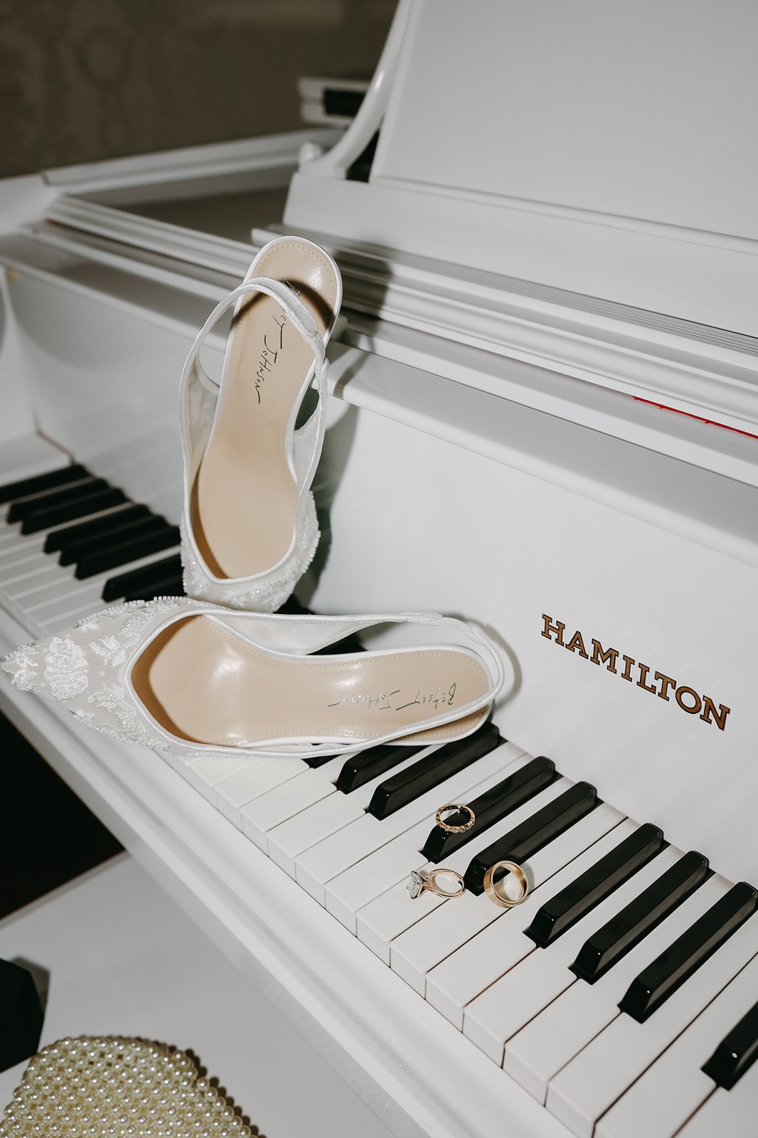A white grand piano with a pair of white wedding shoes, a ring, and a ring with a diamond, placed on the keys. The piano is branded 'Hamilton'.