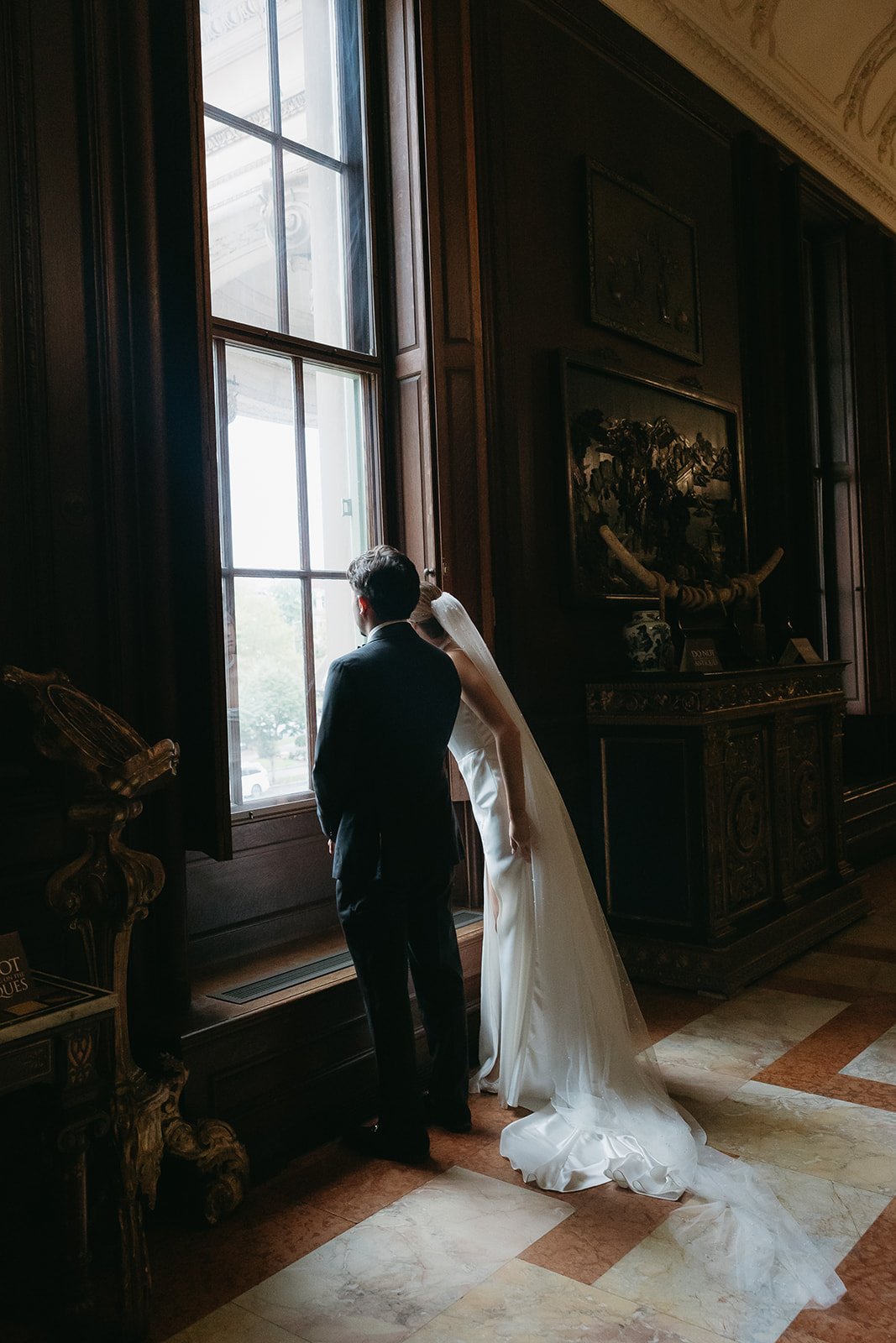 Bride and groom standing by a large window in a historic, ornate room, gazing outside together.