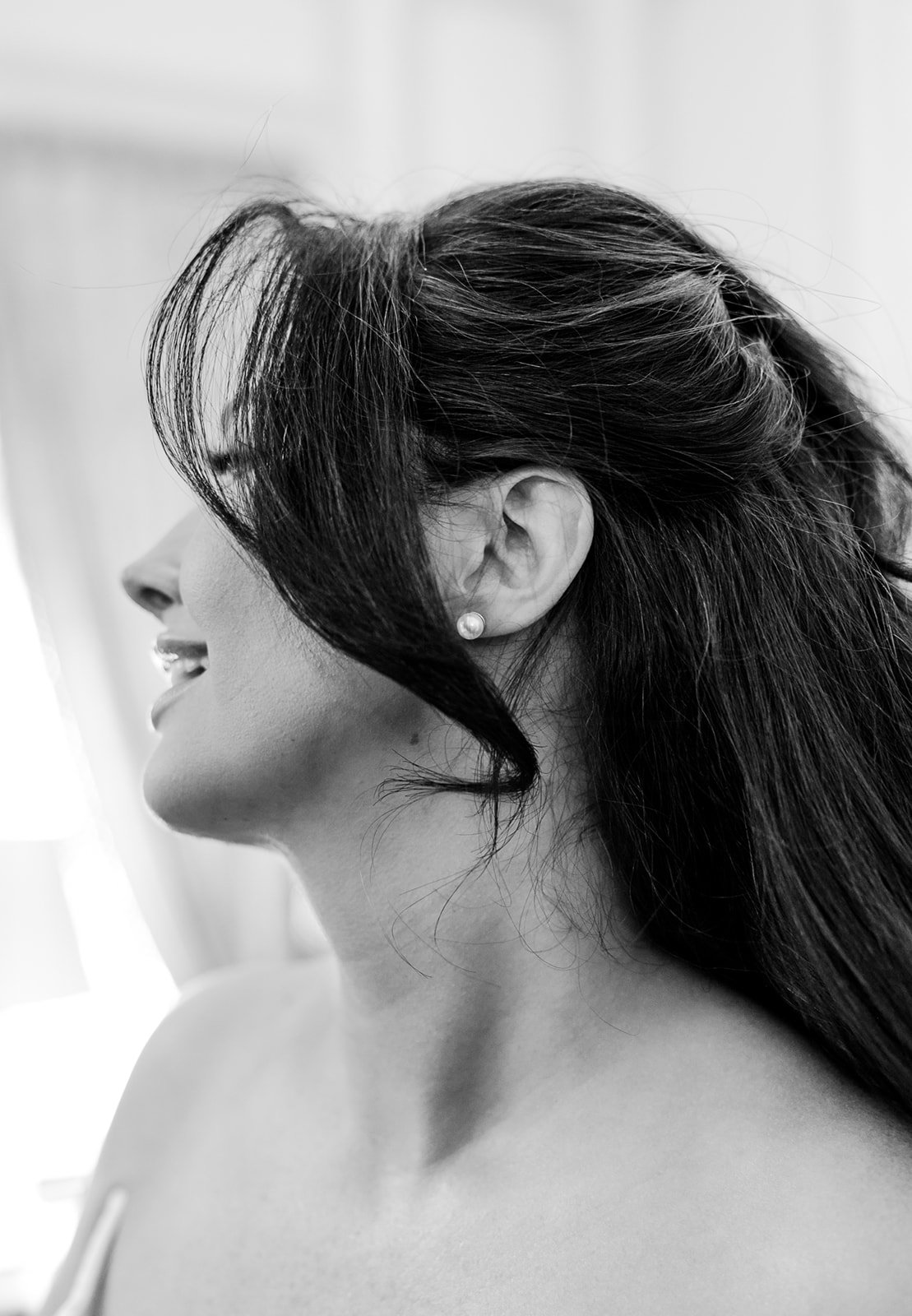 Black and white close-up photo of a woman with long dark hair, pearl earring, sticking out her tongue, with her face turned to the side.
