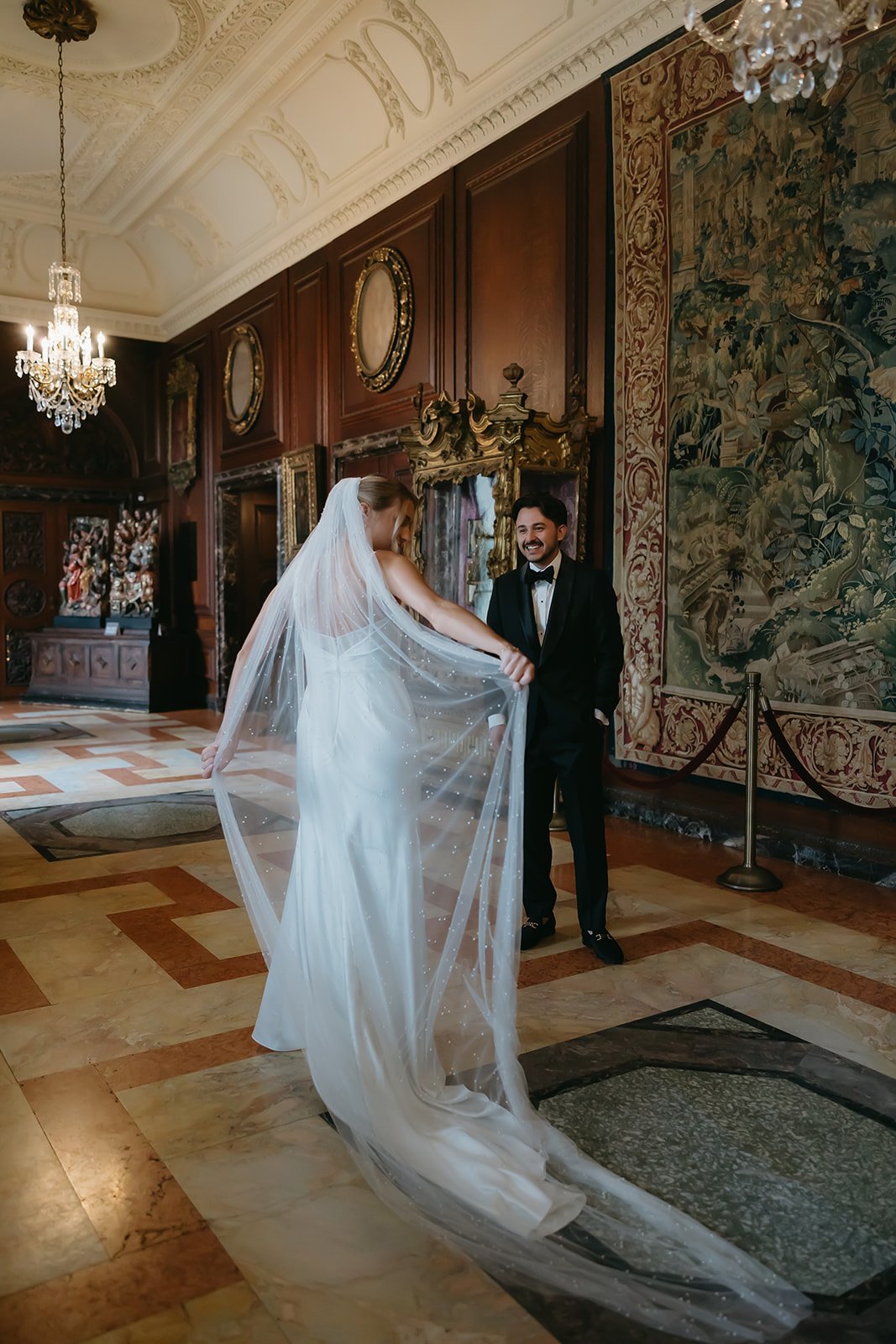 Bride in a wedding gown and veil lifts her dress as groom in a tuxedo smiles indoors at a historic, ornately decorated building.