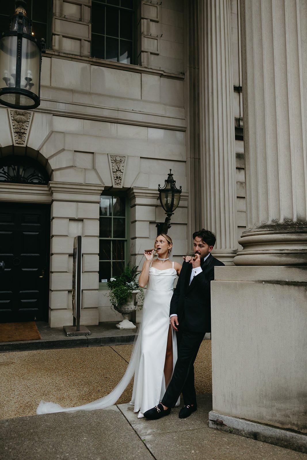 A bride and groom in wedding attire standing outside a grand building, eating popsicles.