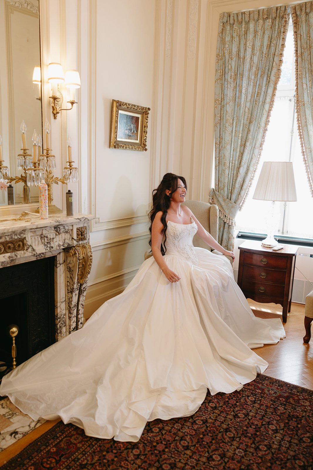 A woman in a wedding dress sitting on a chair in an elegant room with cream-colored walls, ornate curtains, and vintage furniture, smiling and enjoying the moment.