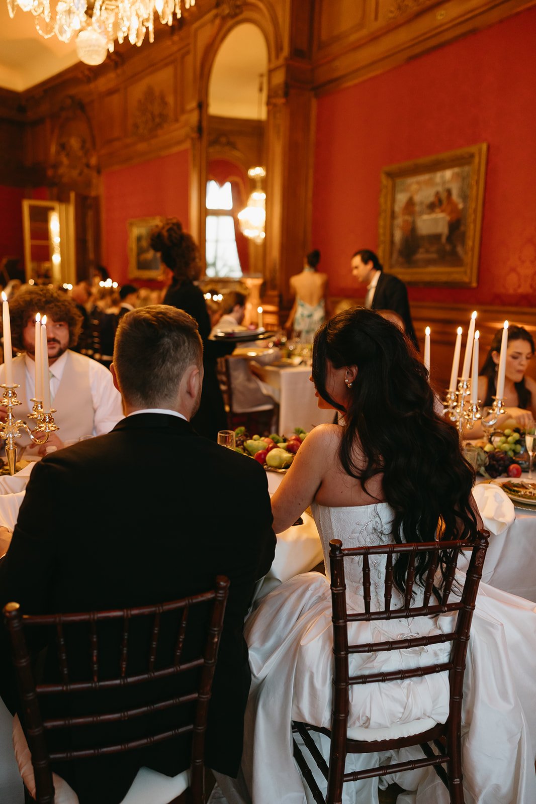A bride and groom sitting at a formal dinner table during their wedding reception in an elegant, wood-paneled room with red wallpaper, paintings, and a chandelier.