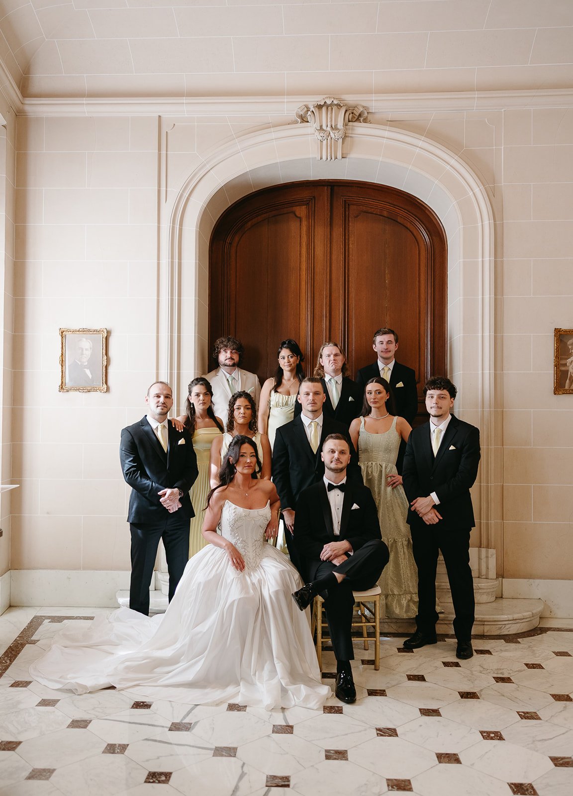 Group of wedding guests dressed in formal attire, with the bride in a white gown and the groom in a tuxedo, posing in a grand hall with wooden doors and marble floors.