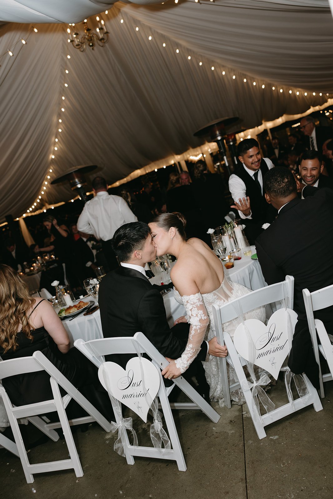 Couple in wedding attire kissing at their wedding reception, decorated with heart and 'Just married' signs on chairs, under a draped tent with string lights.