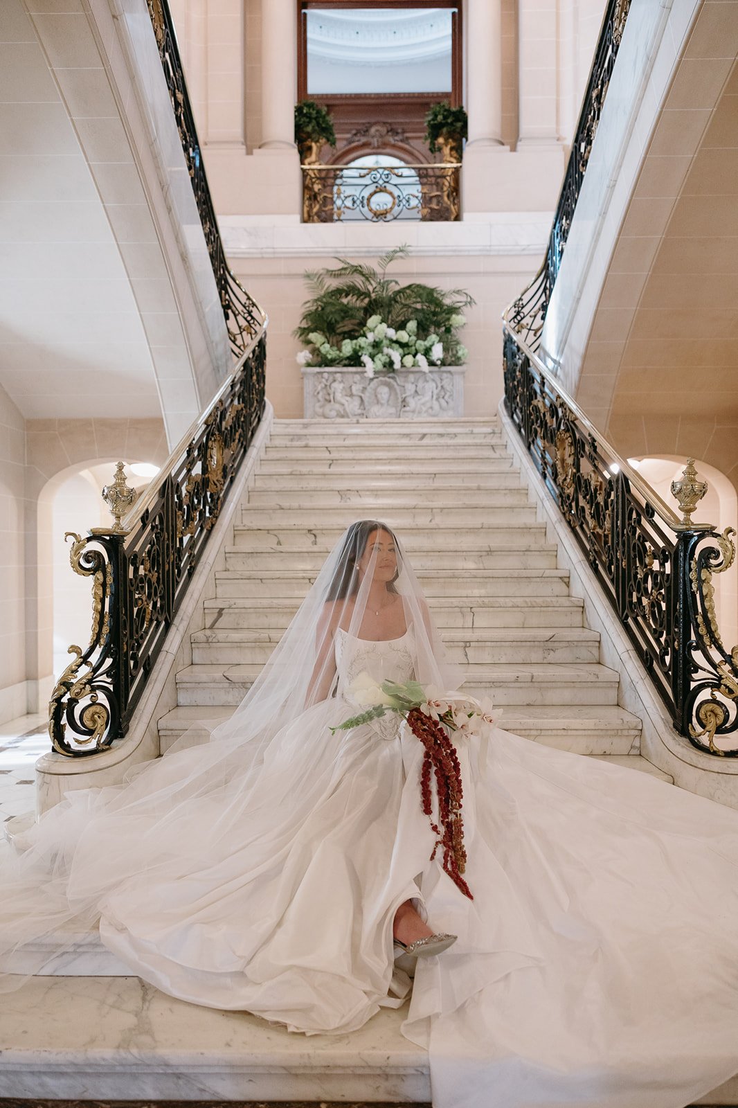 Bride in a white wedding gown sitting on a grand marble staircase with ornate black and gold railings, holding a bouquet with red and white flowers, in an elegant interior setting.