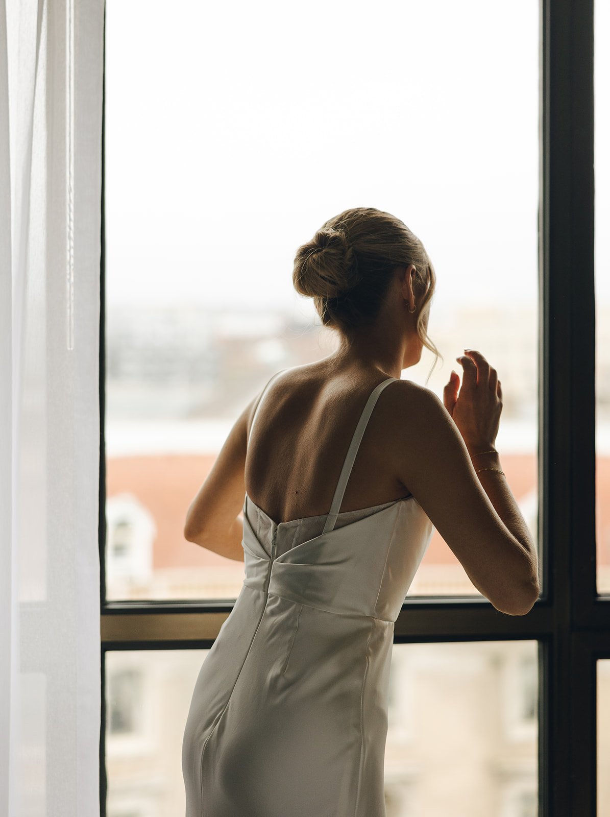 A woman in a white dress standing by a large window, looking outside and touching the window with her hand, with her hair styled in an elegant updo.