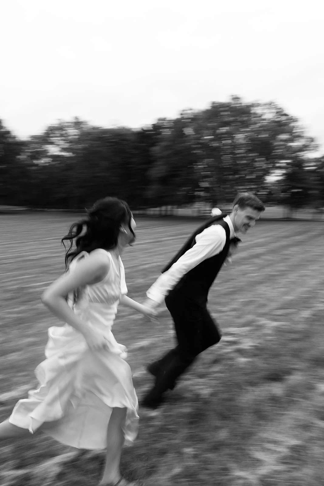 A bride in a white wedding gown and a groom in a tuxedo sitting on a vintage red floral sofa in a decorated room with large windows, yellow curtains, a chandelier, and framed religious artwork on the wall.
